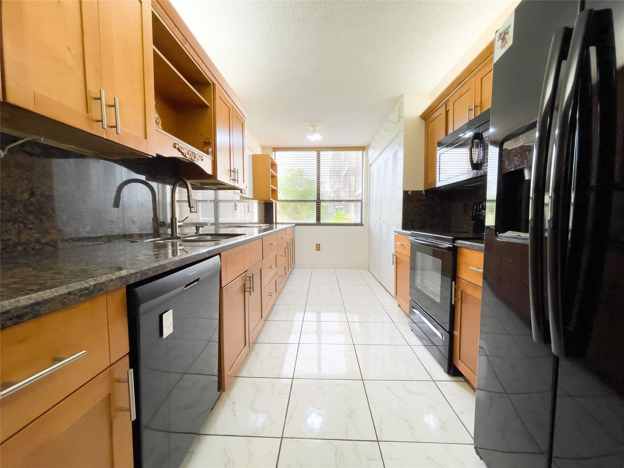 Galley kitchen with warm wood cabinets, dark counters, black appliances, and a large end window.