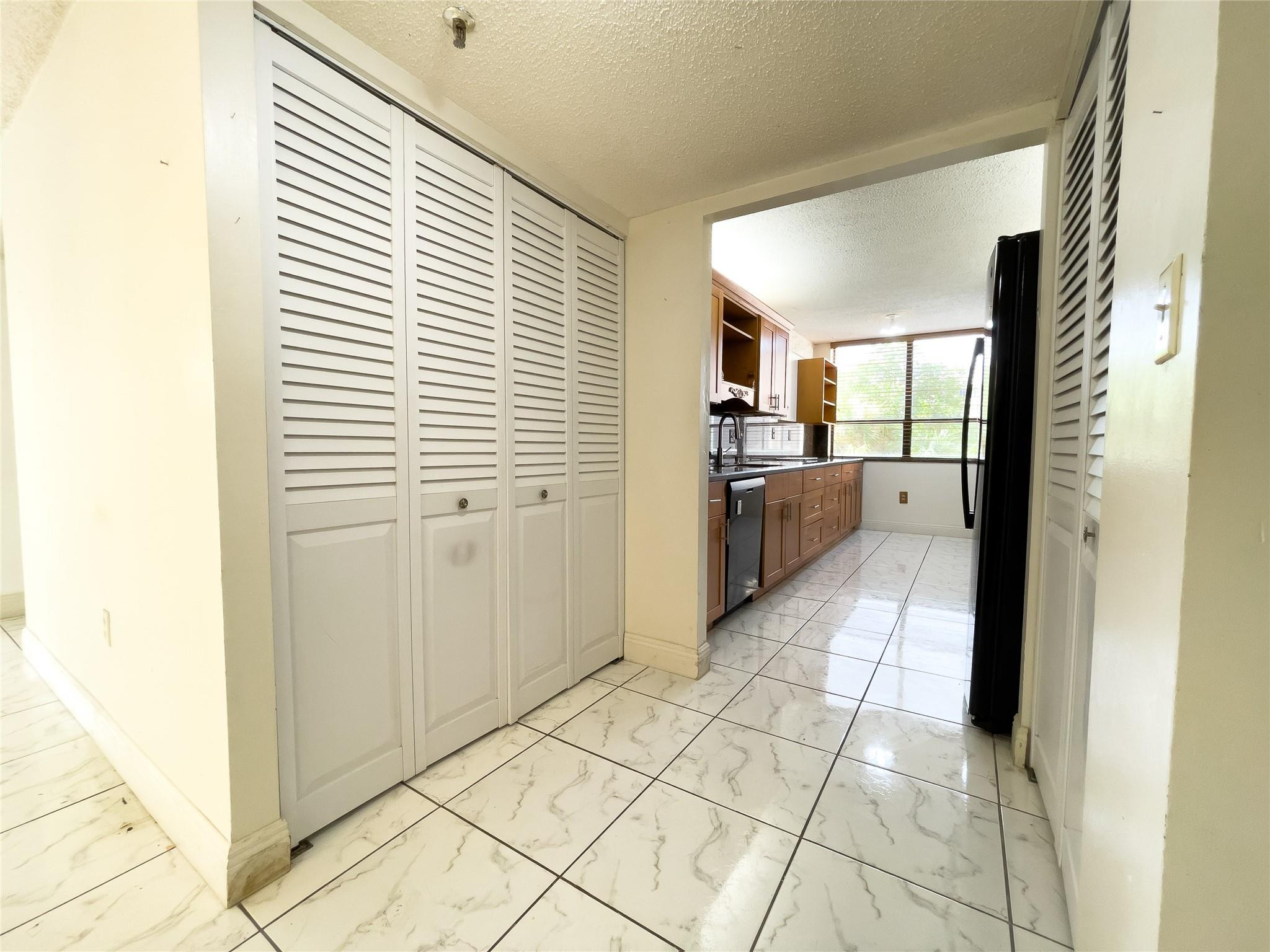 Condo hallway with white louvered closets and marble-pattern tile leading to a wood-cabinet kitchen.