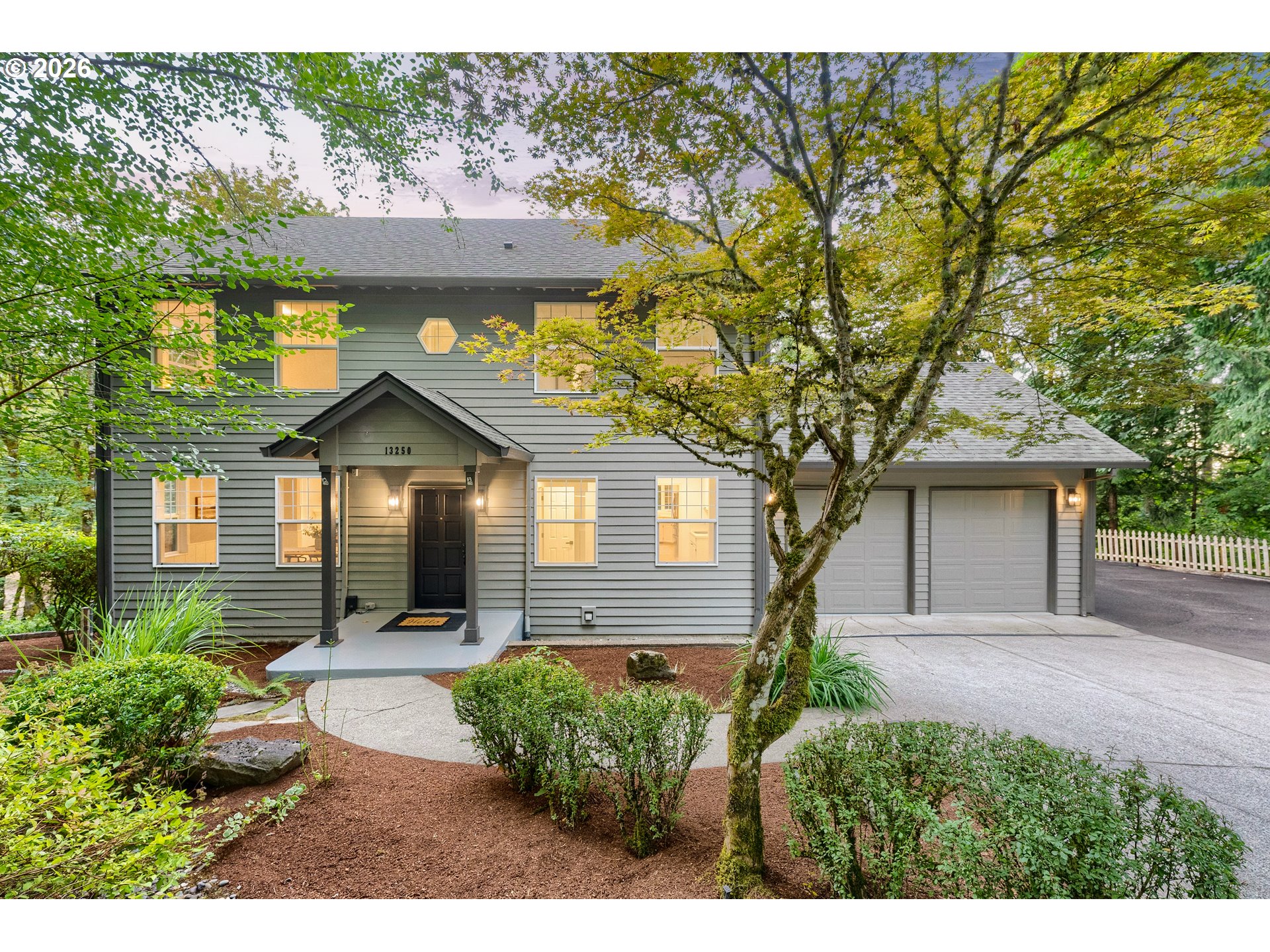 Gray two-story house with a covered entrance, two-car garage, and mature trees in the landscaped yard.