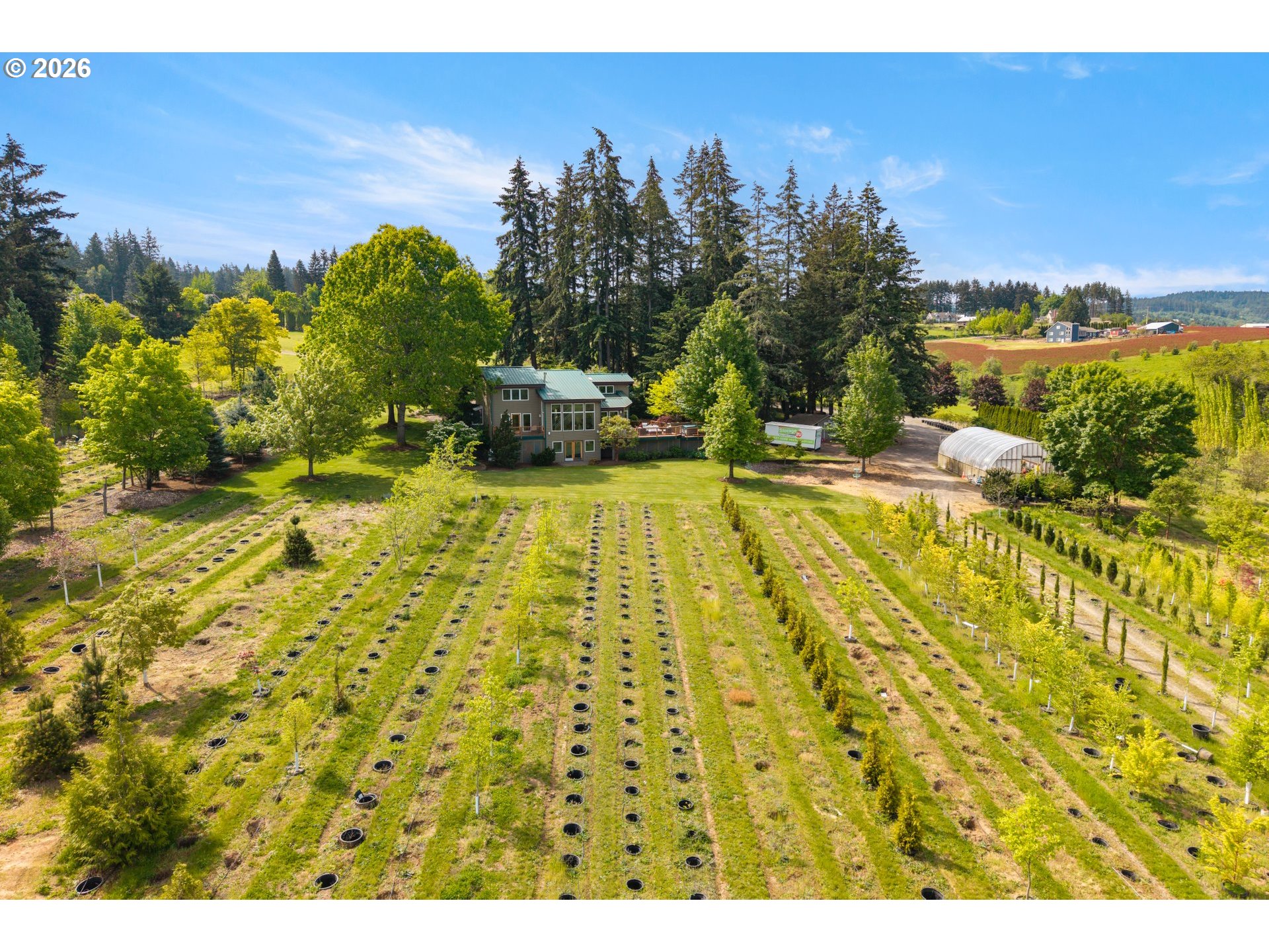 Aerial view of a rural home surrounded by rows of plantings, mature trees, and a greenhouse.