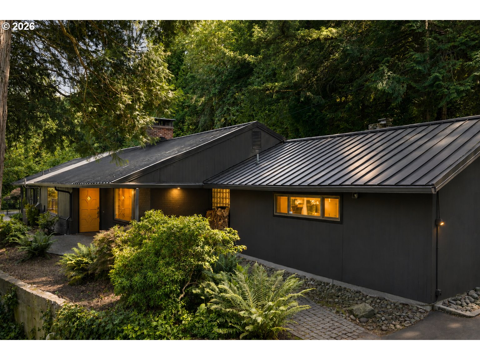 Contemporary dark house with a metal roof, glowing windows, and lush garden landscaping.
