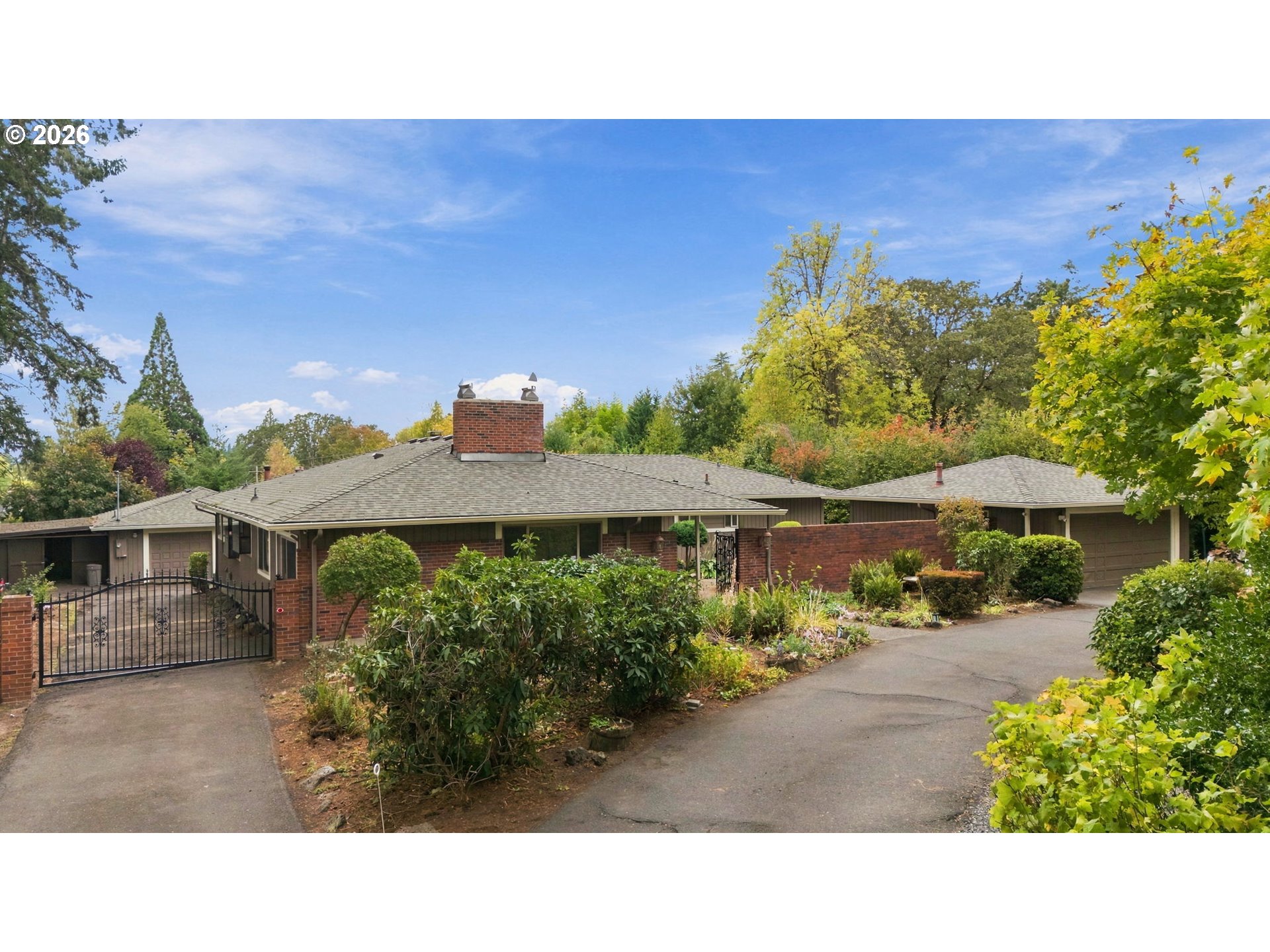 Single-story brick home with a gated driveway, attached garages, and mature landscaped gardens.