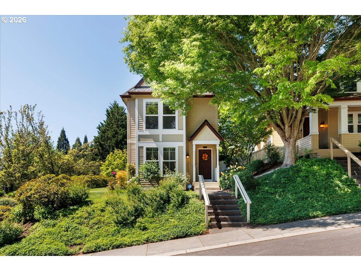 Two-story beige house with a dark front door, white-railed entry stairs, and a mature tree in the garden.