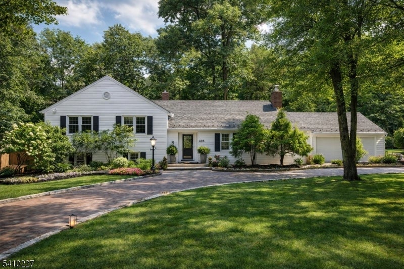 White single-story home with black shutters, attached garage, and curved brick driveway beneath mature trees.