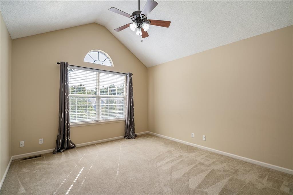 Empty carpeted bedroom with a vaulted ceiling, ceiling fan, and arched window above the curtained main window.