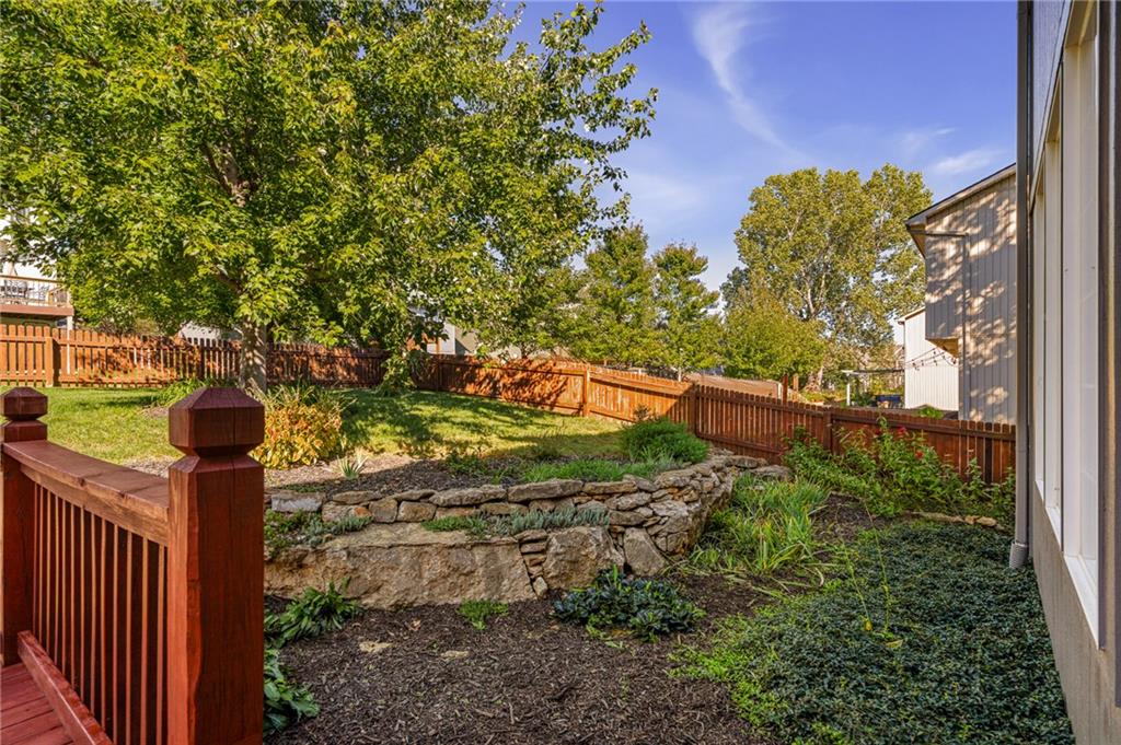 Landscaped backyard with a wooden deck, tiered stone walls, garden beds, and mature trees.