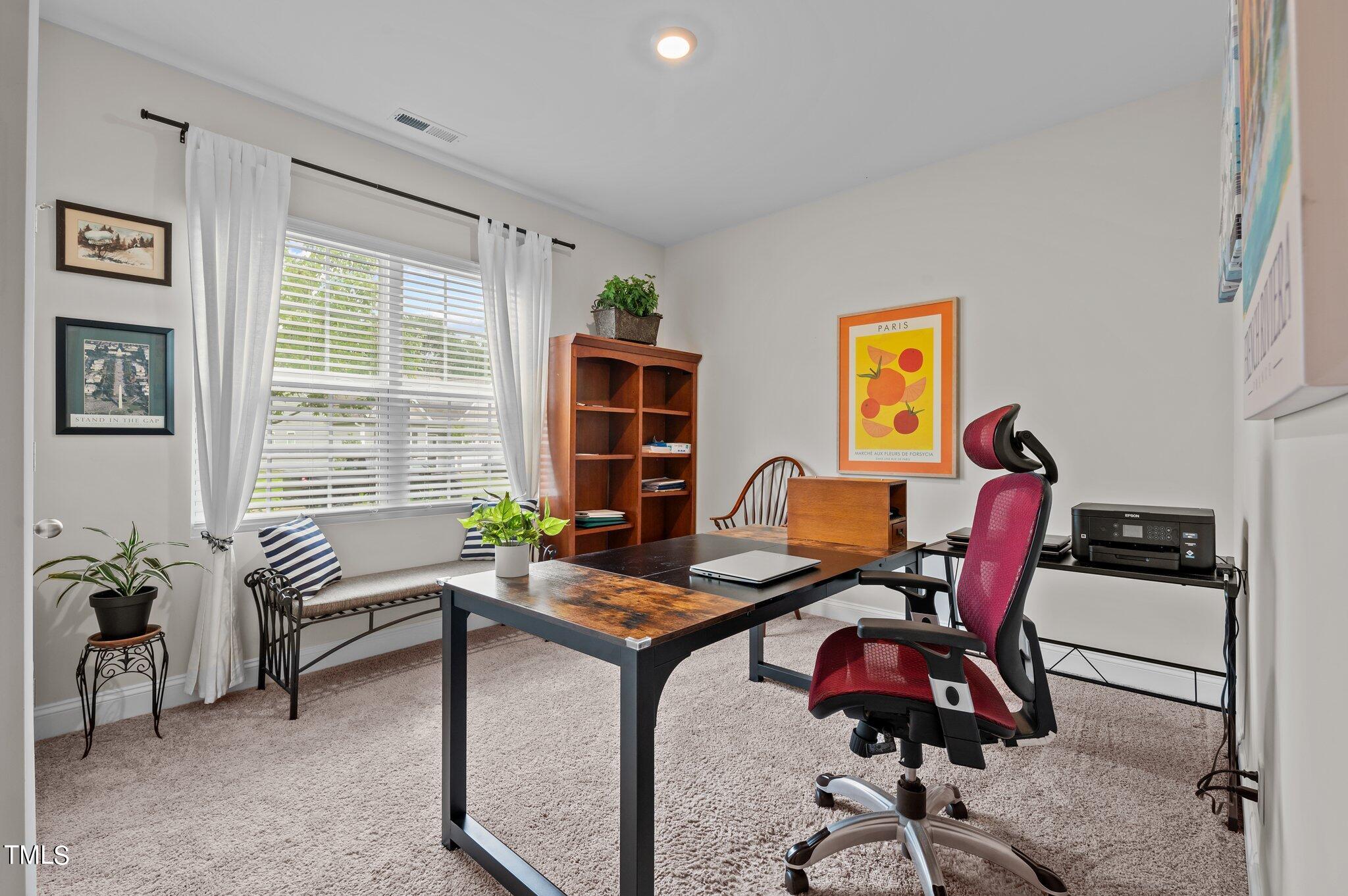 Furnished home office with a large curtained window, window seat, wooden bookcase, and desk.