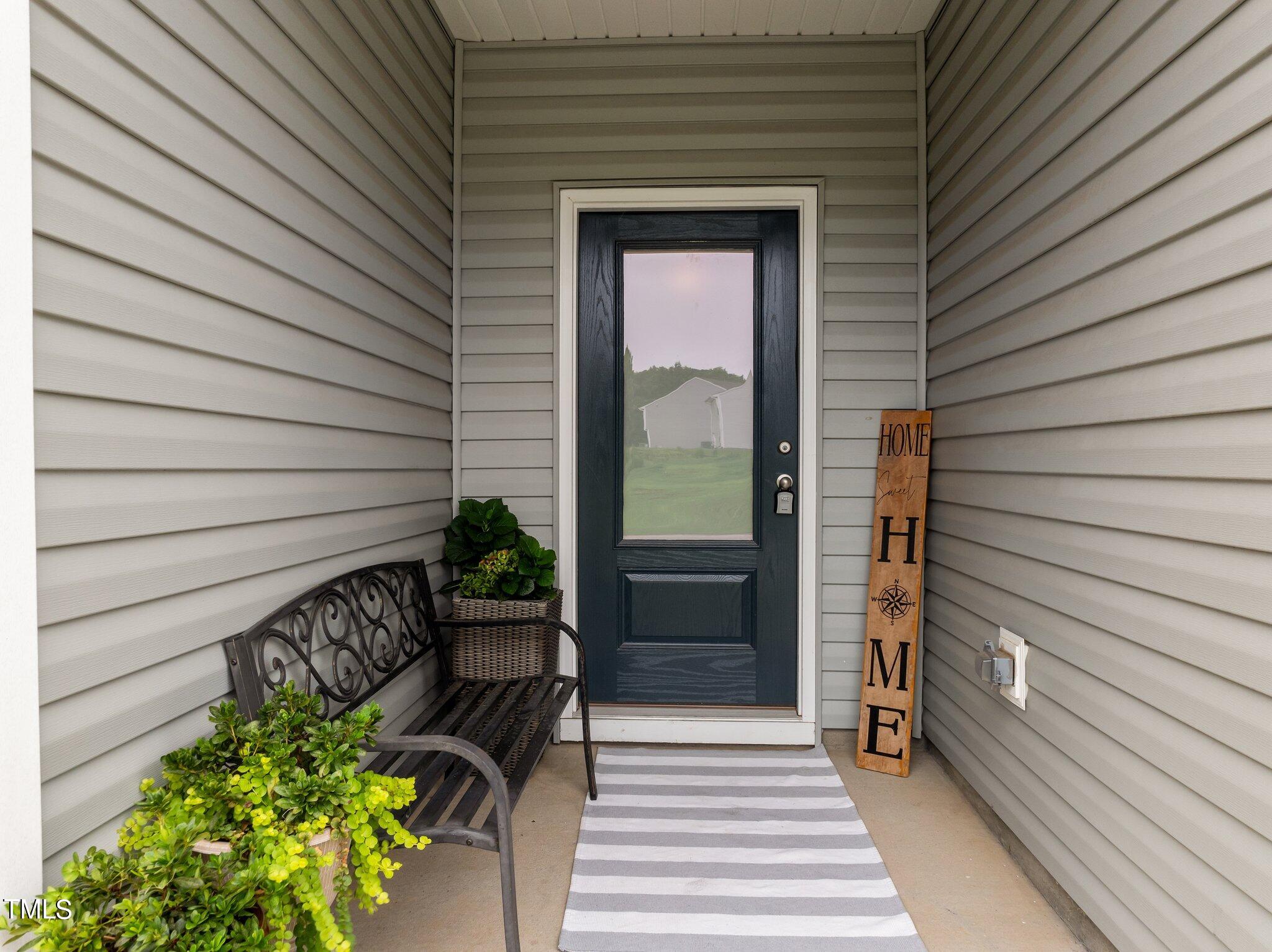 Covered home entry with a dark glass-panel door, metal bench, potted plants, and striped doormat.