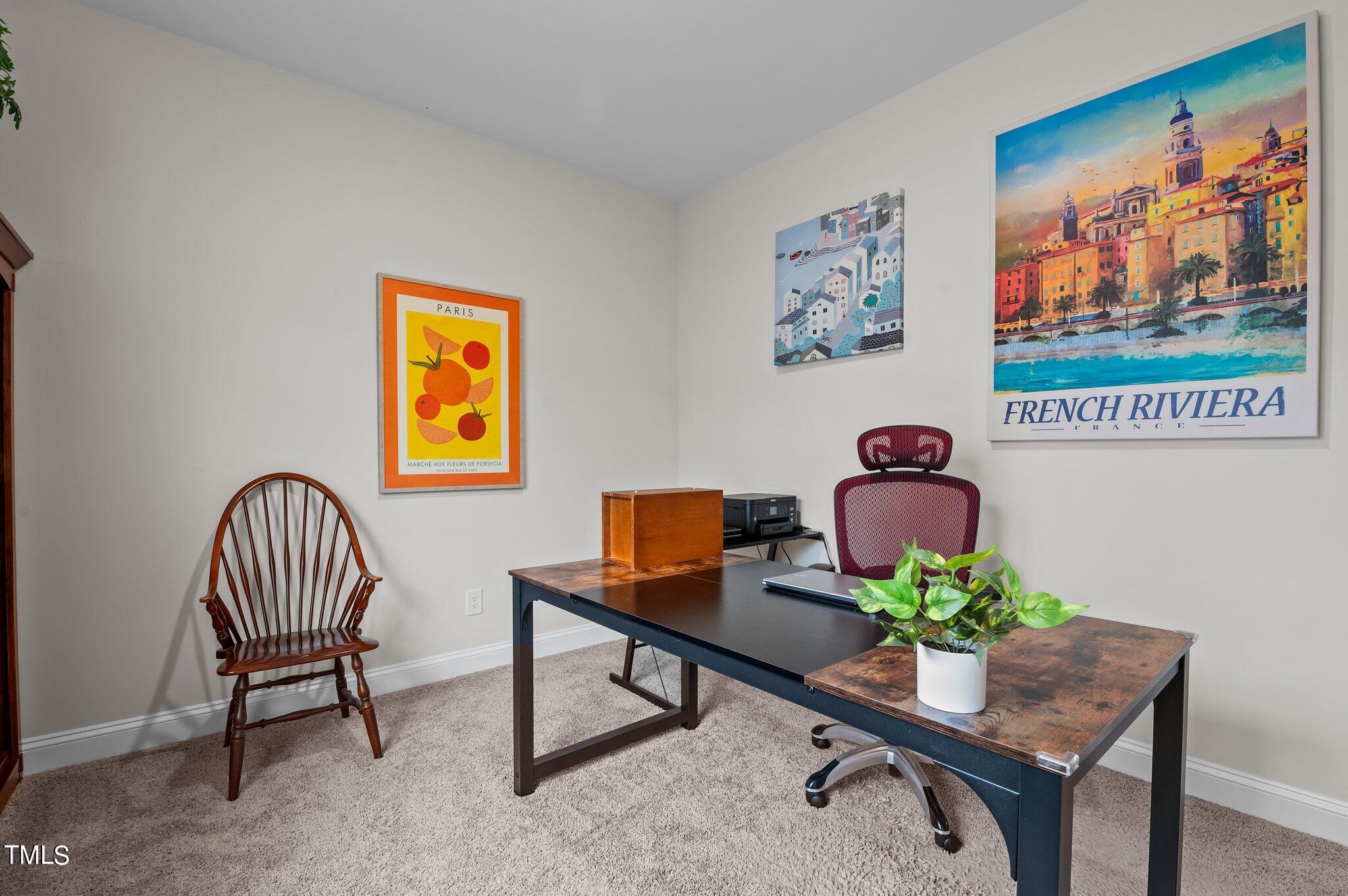 Furnished home office with a dark desk, burgundy rolling chair, wooden chair, and colorful travel posters.