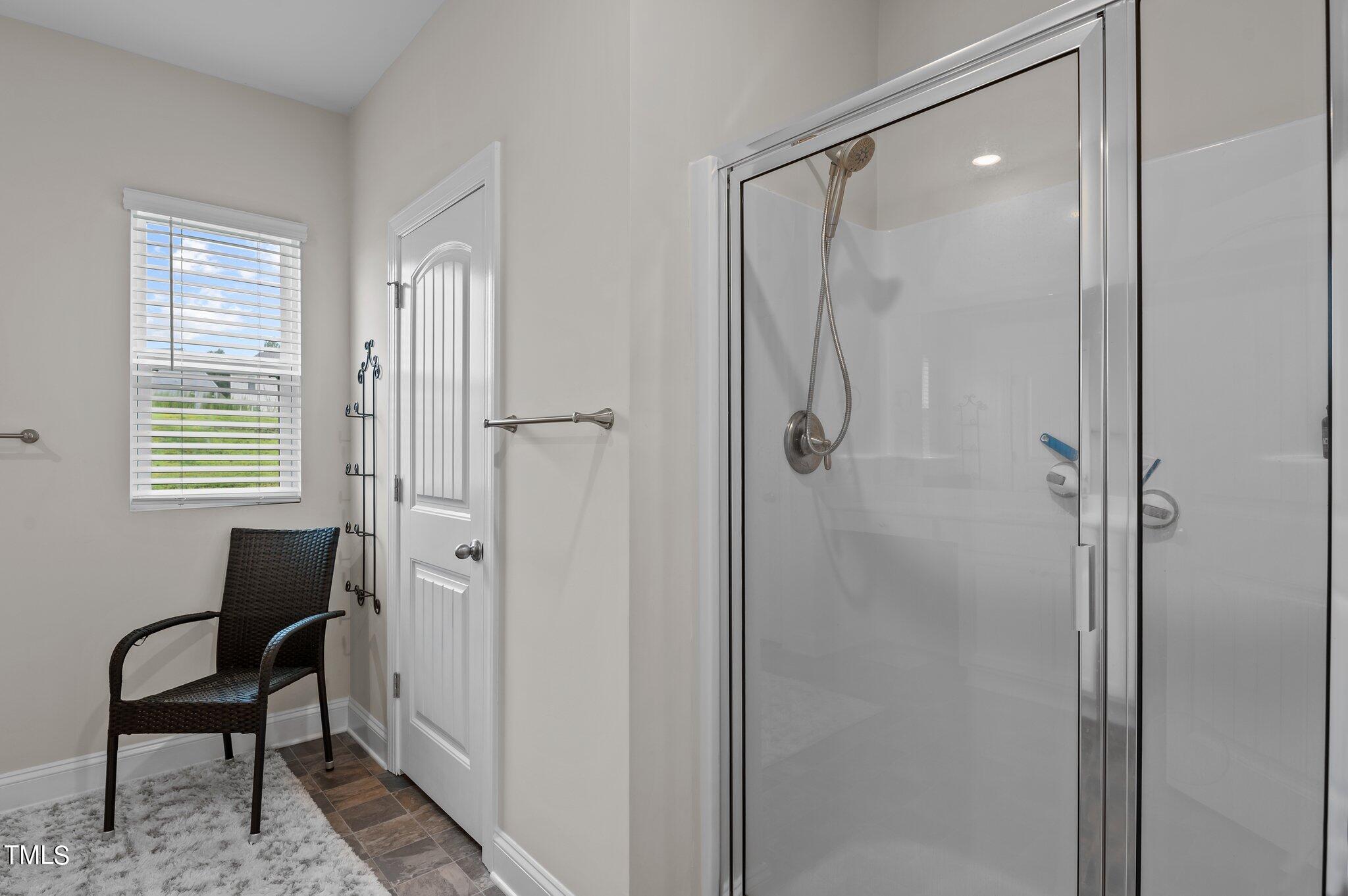 Bathroom with a glass shower, tiled floor, and wicker chair beside a window.