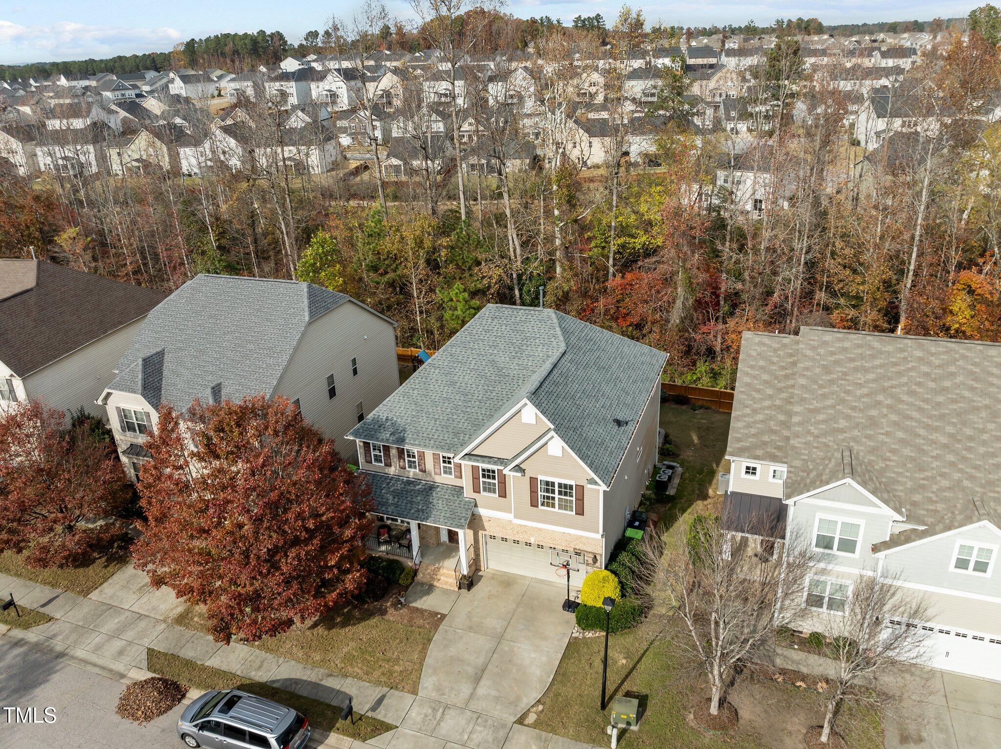 Aerial view of a beige two-story house with an attached garage, driveway basketball hoop, and wooded backdrop.
