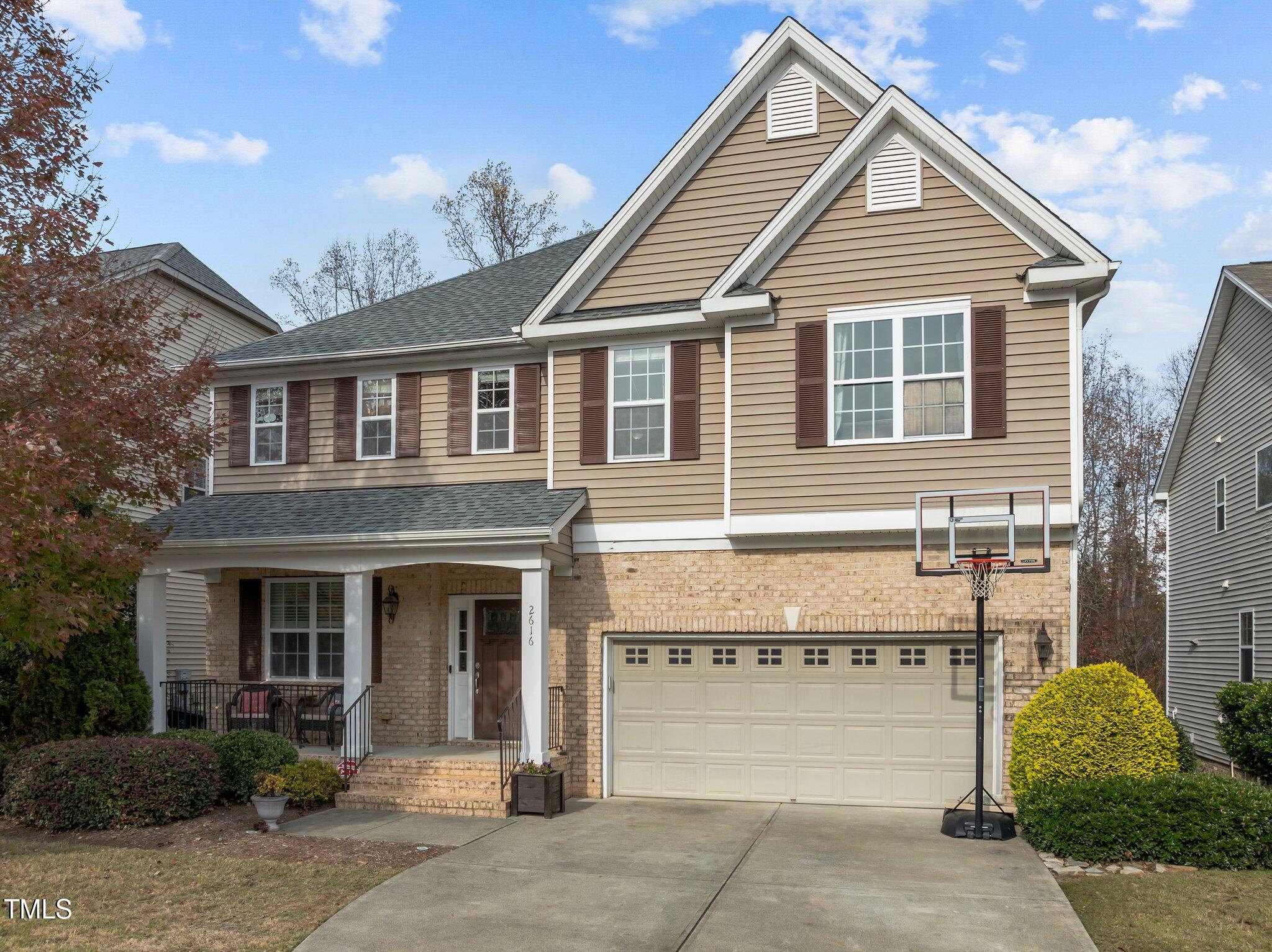 Two-story beige house with a brick base, covered porch, double garage, and driveway basketball hoop.