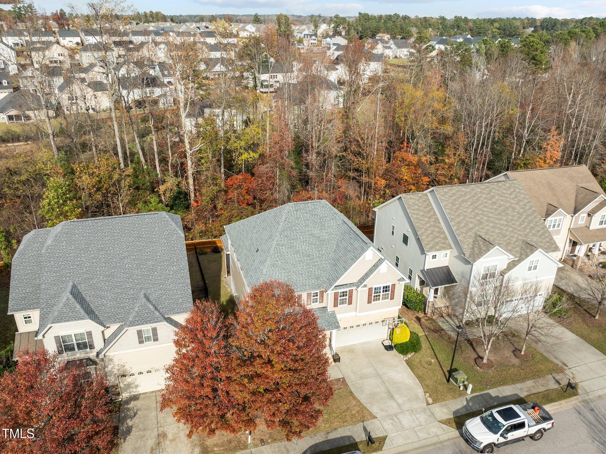 Aerial view of a tan home beside autumn woods and neighboring houses.