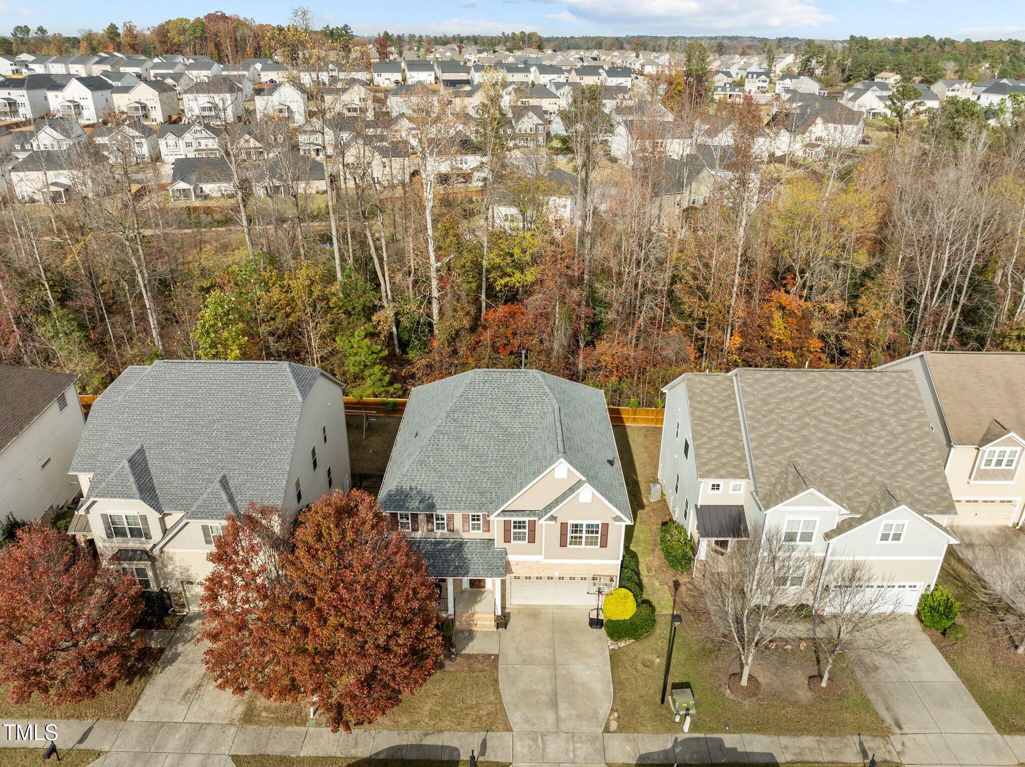 Aerial view of a tan two-story house beside an autumn-colored wooded buffer and neighborhood homes.