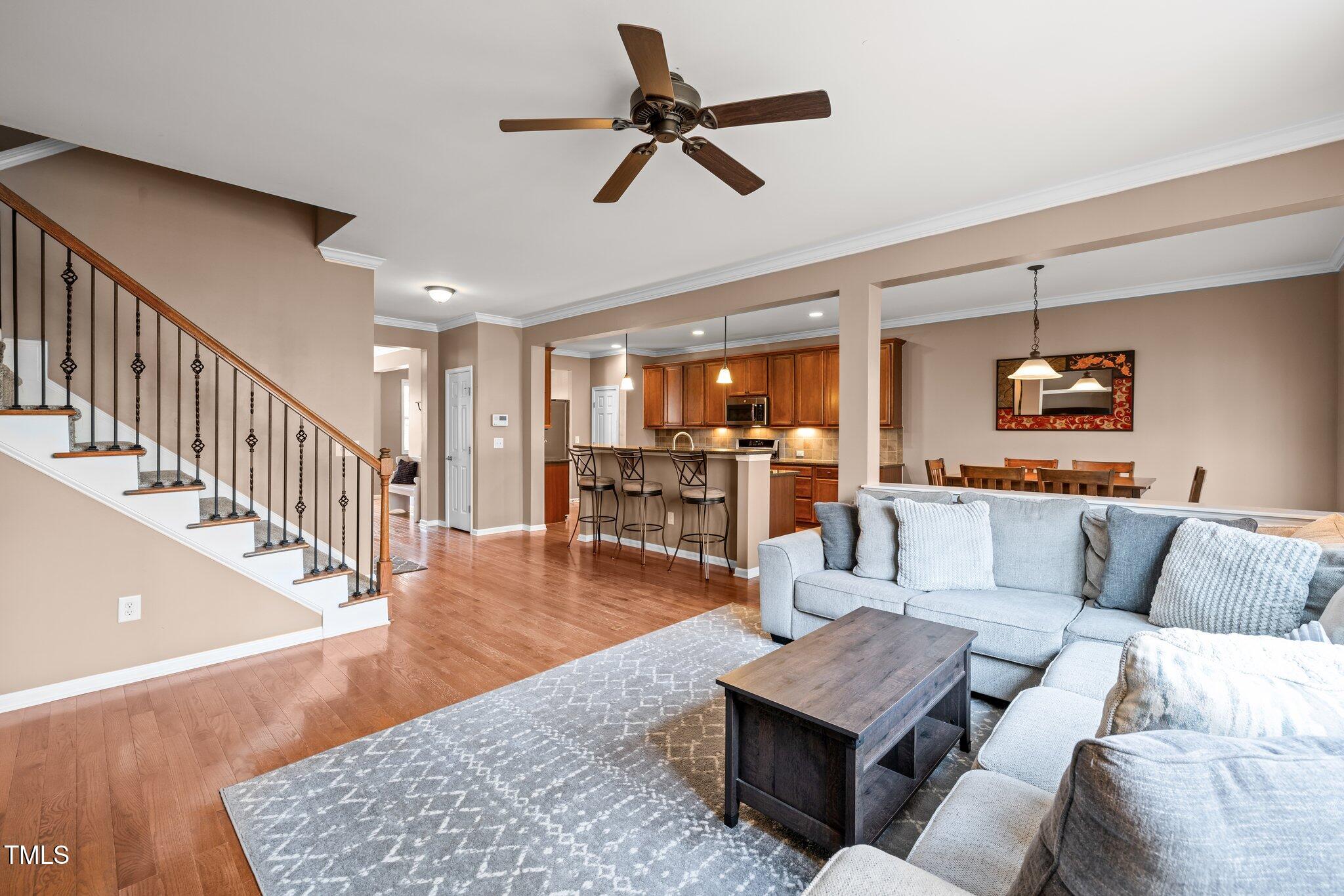 Open-plan living room with gray sectional, hardwood floors, staircase, and adjoining kitchen and dining area.