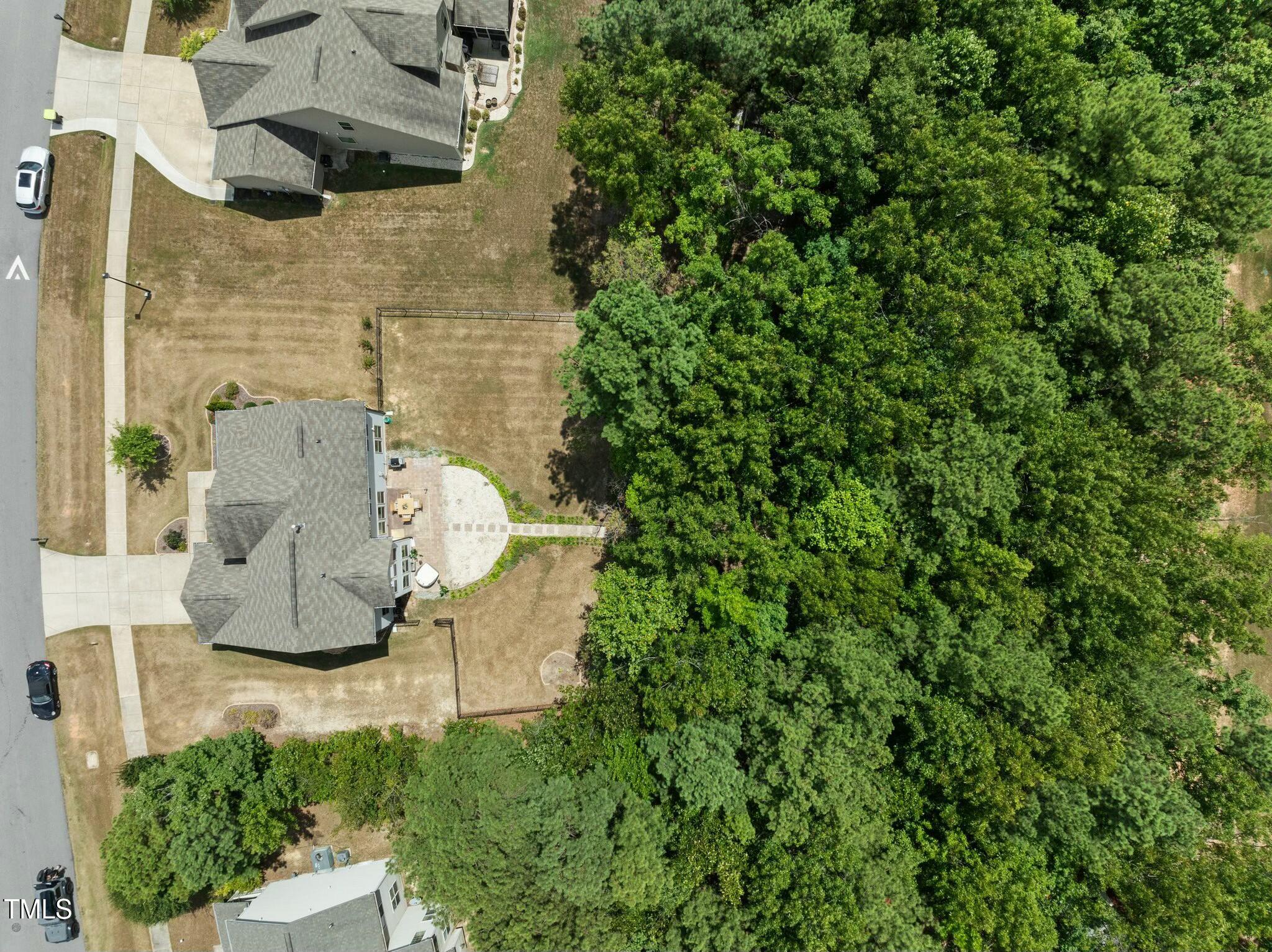 Aerial view of a home and yard beside dense woods, with a paved patio and fenced sections.