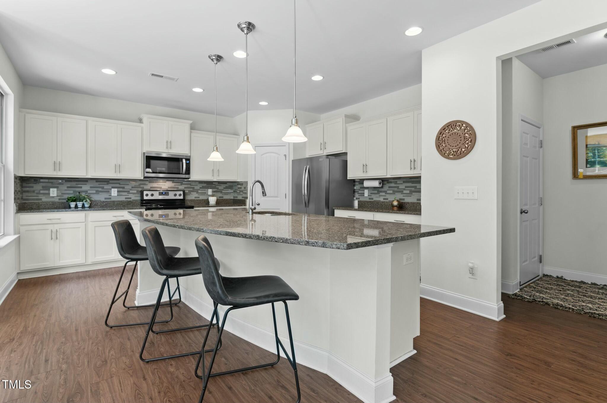 Kitchen with white cabinets, stainless appliances, and a large granite island with three black stools.