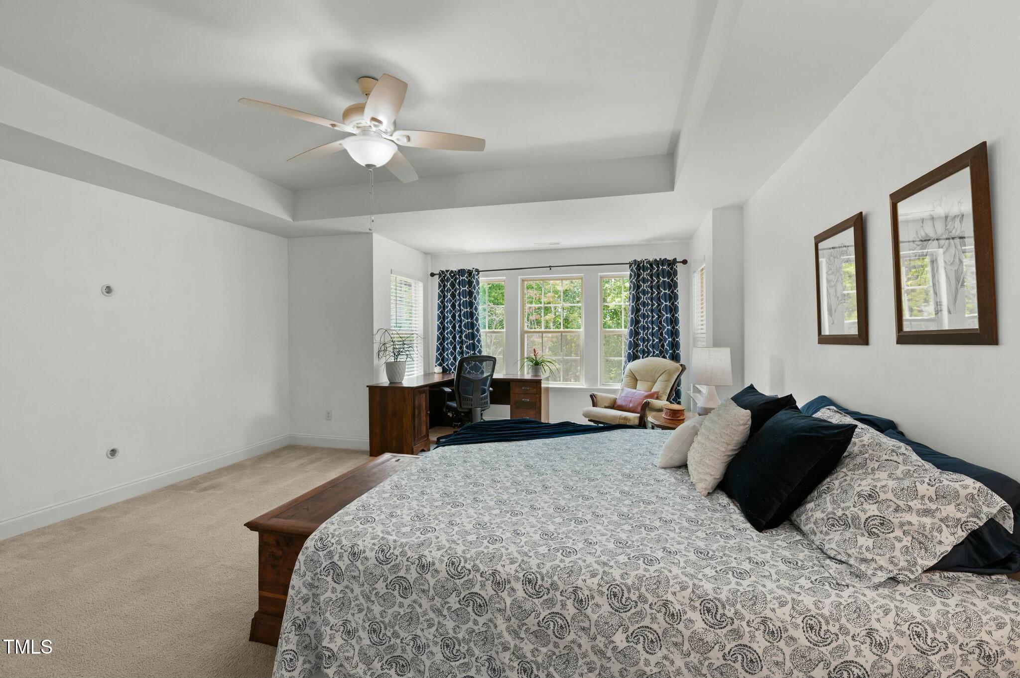 Furnished bedroom with a large patterned bed, tray ceiling, and desk beside curtained windows.