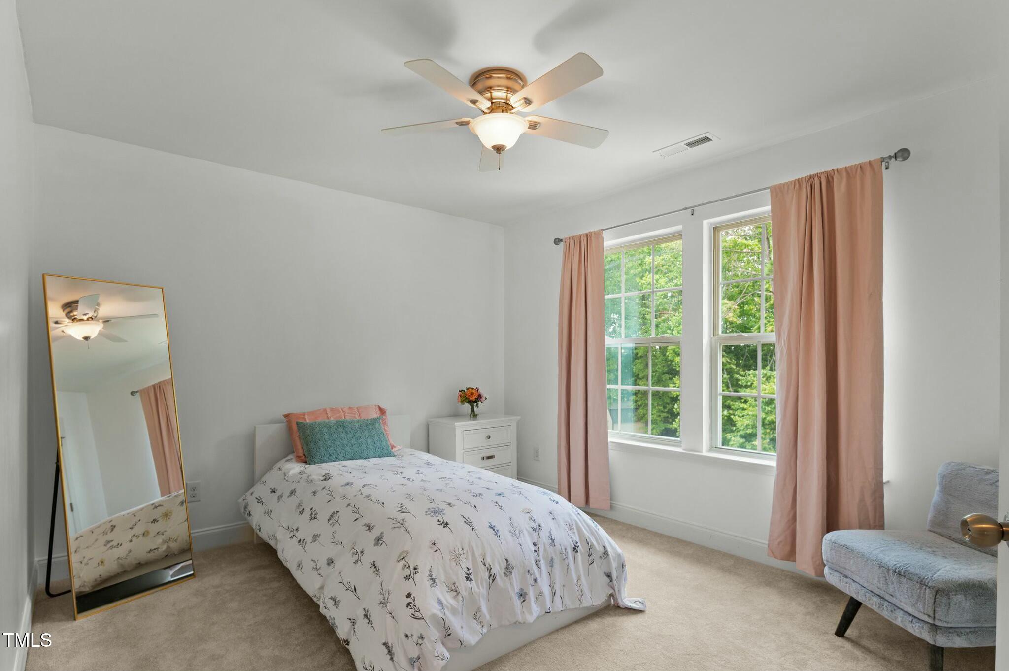 Furnished bedroom with a floral bedspread, blush curtains, and two windows overlooking green trees.