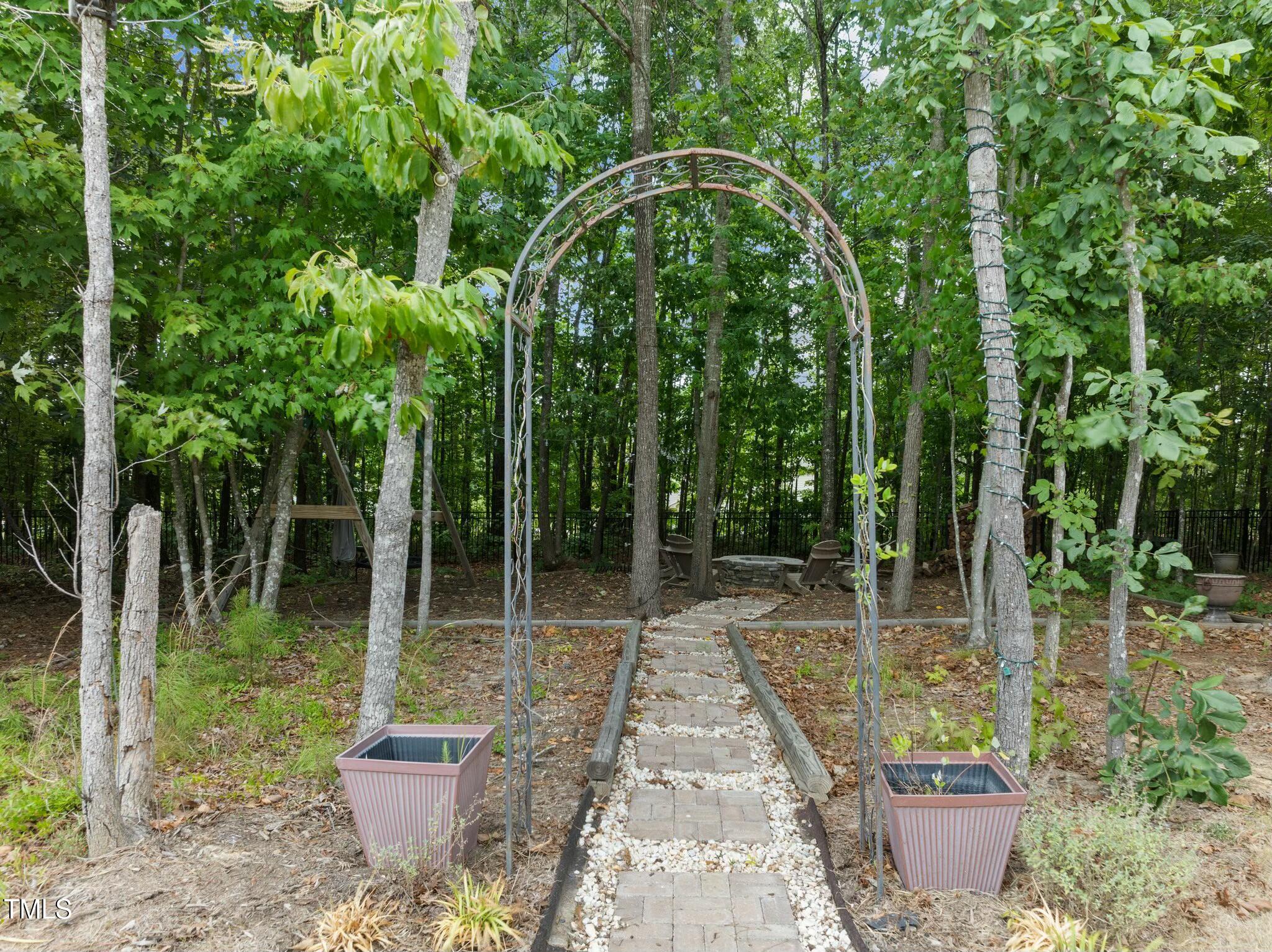Wooded backyard path passes beneath a metal garden arch toward a fire pit seating area.