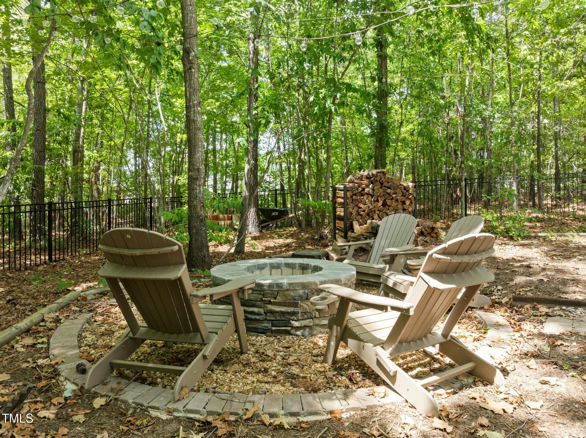 Stone fire pit surrounded by Adirondack chairs beneath string lights in a wooded yard.