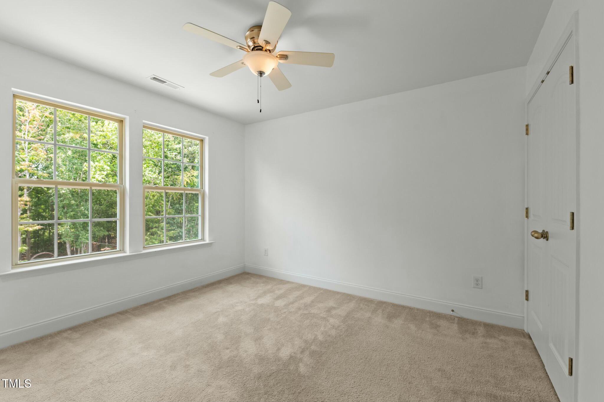 Empty carpeted bedroom with two windows overlooking trees and a ceiling fan.