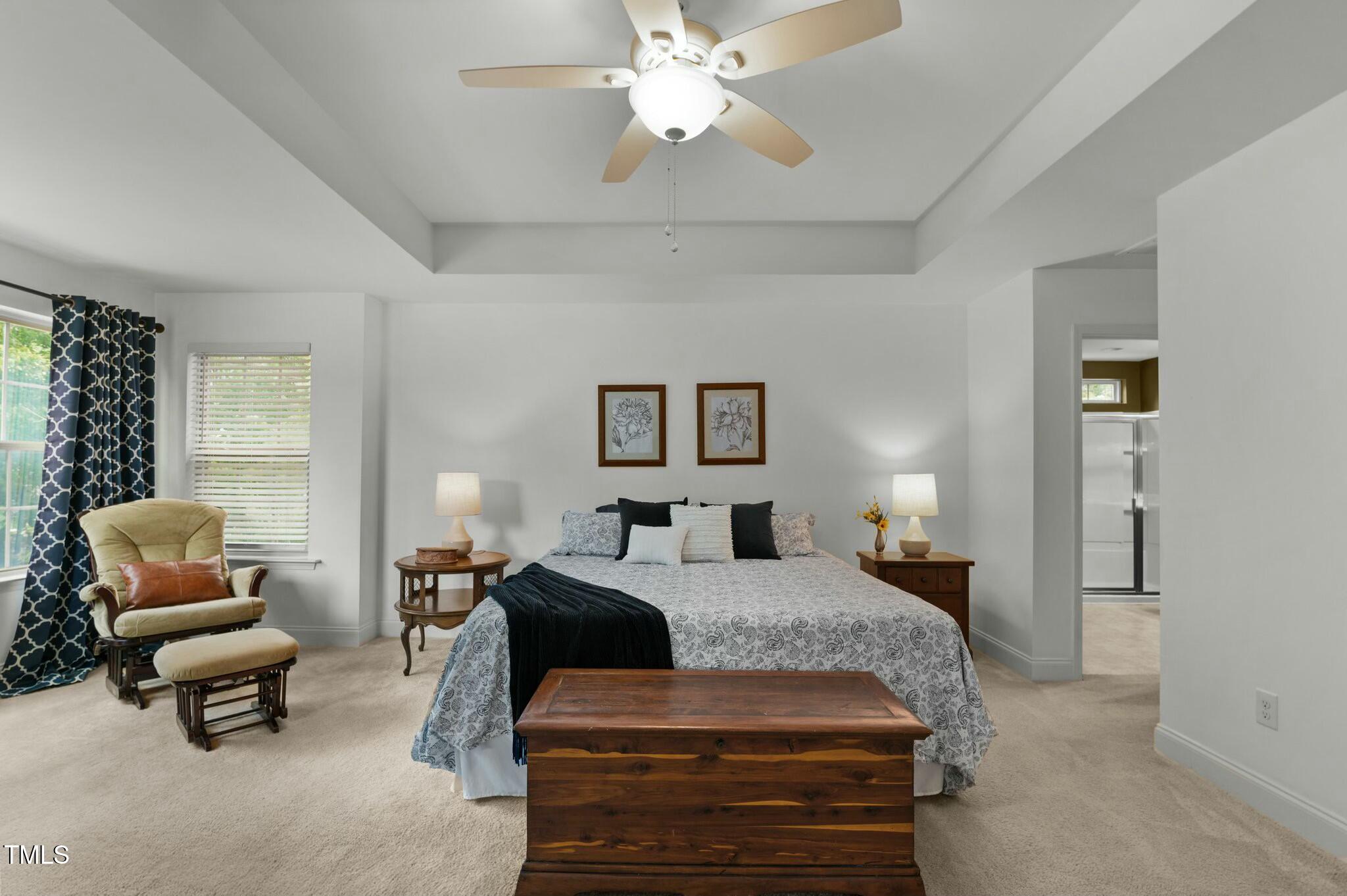 Staged bedroom with a large bed, wooden storage bench, and chair beside curtained windows.