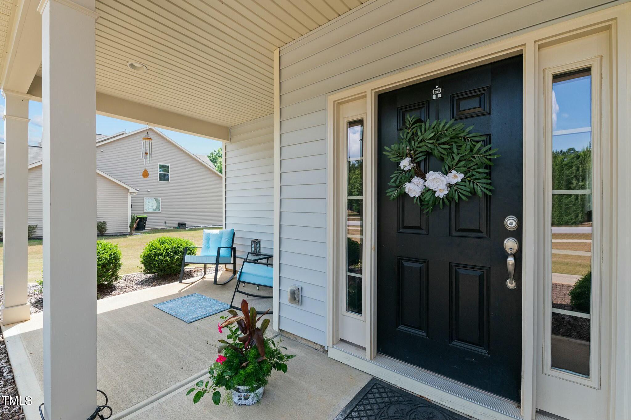 Covered front porch with a black door, floral wreath, blue chairs, and potted flowers.