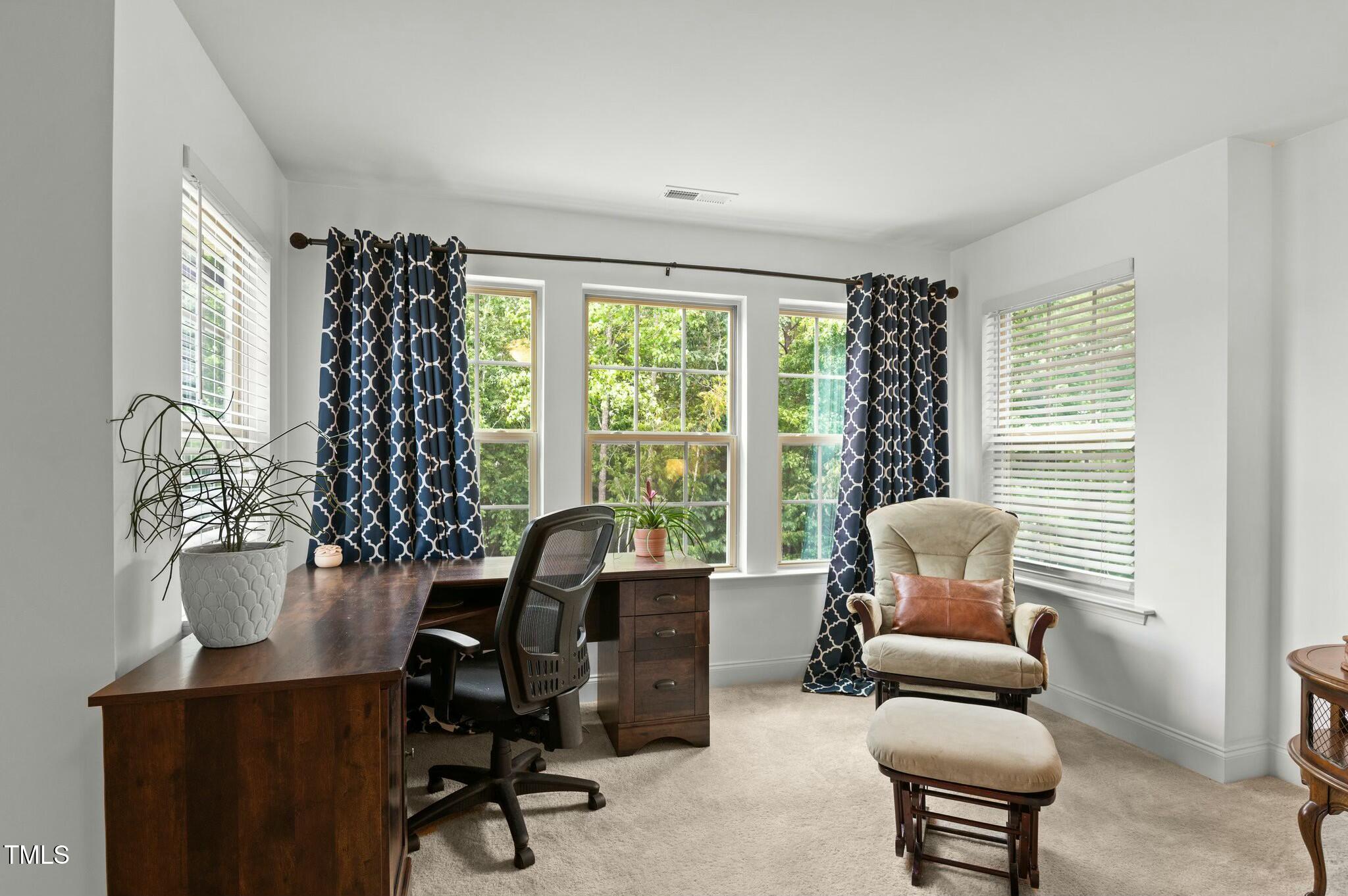 Furnished home office with a dark wood desk, navy patterned curtains, and a beige armchair by the windows.