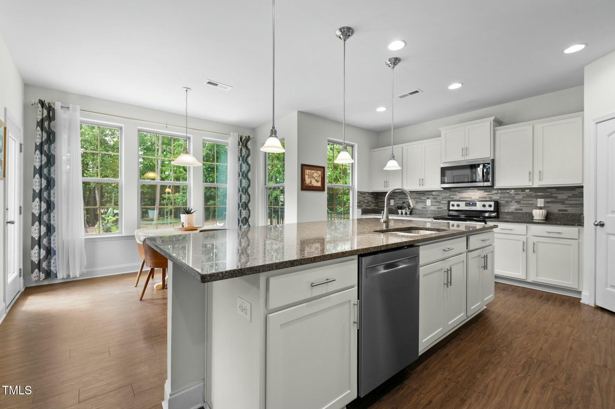 Bright kitchen with a large granite island, white cabinets, stainless steel appliances, and windows overlooking trees.