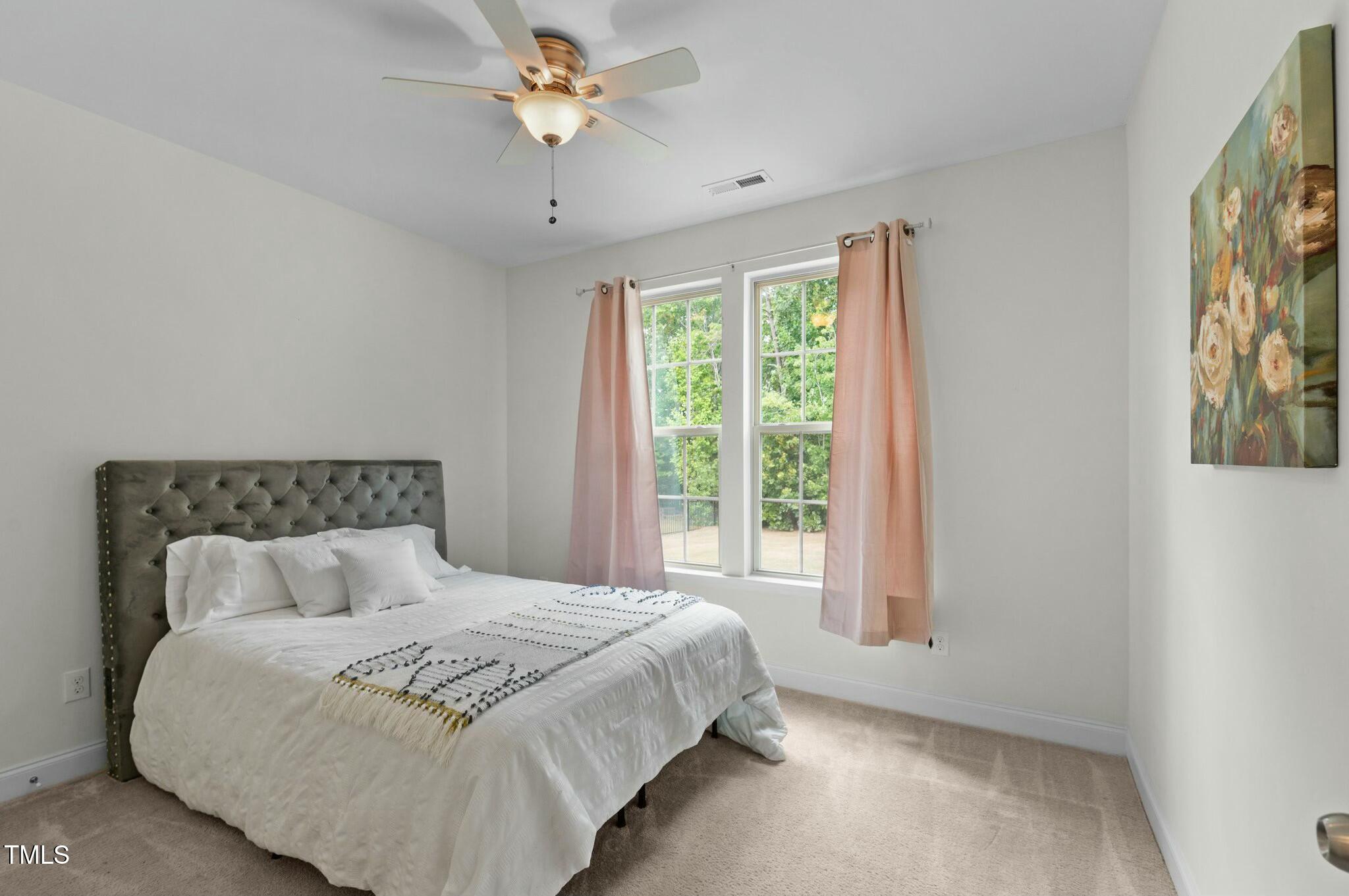 Furnished bedroom with a white bed, tufted gray headboard, and pink curtains beside two windows.