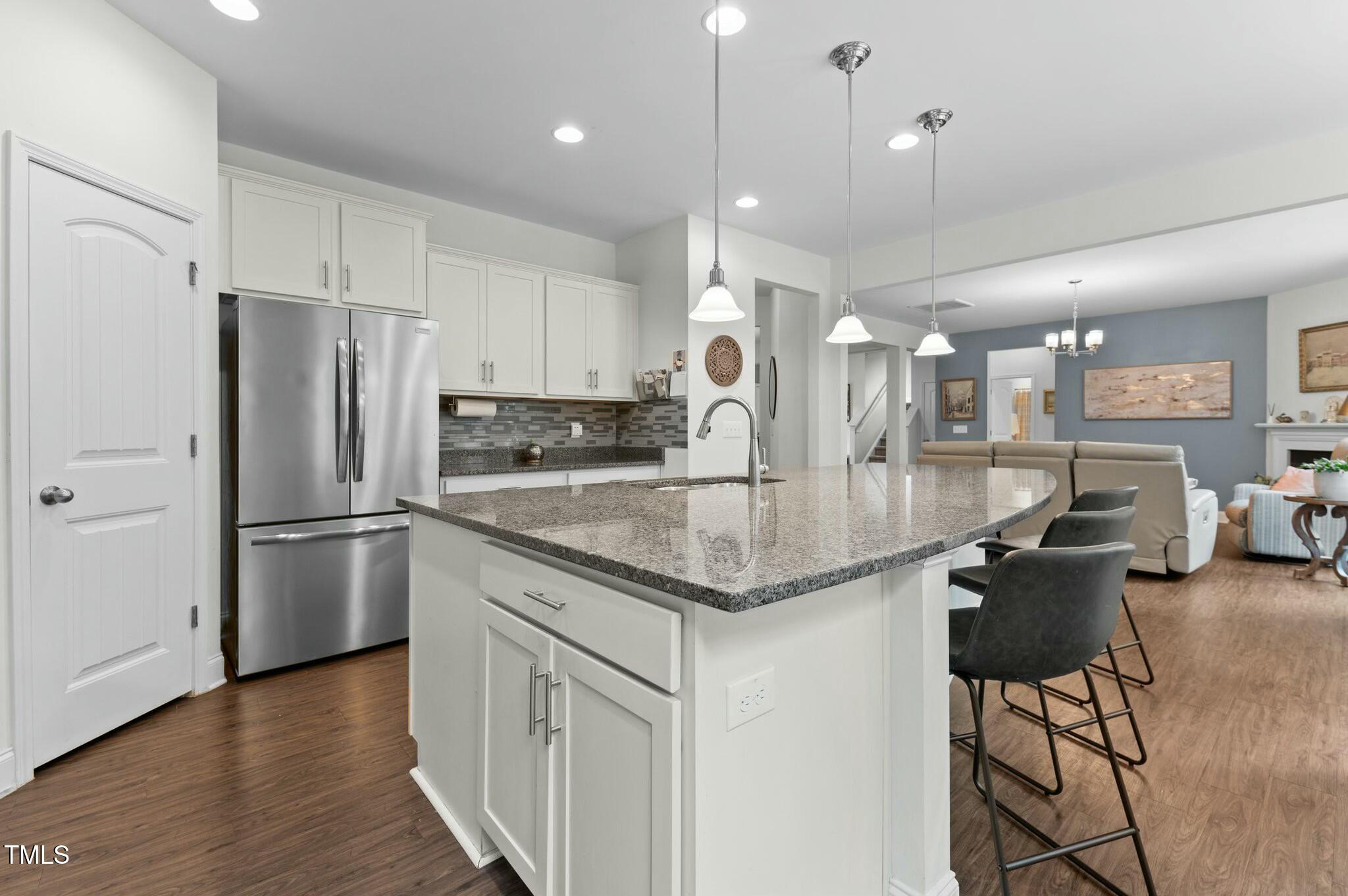 Open-plan kitchen with a large granite island, white cabinets, and stainless appliances beside a furnished living room.