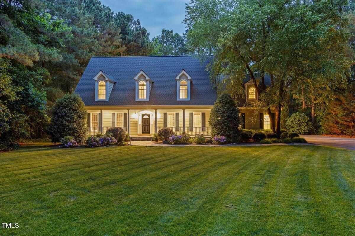 Two-story home with illuminated dormers and a covered porch, surrounded by mature trees.