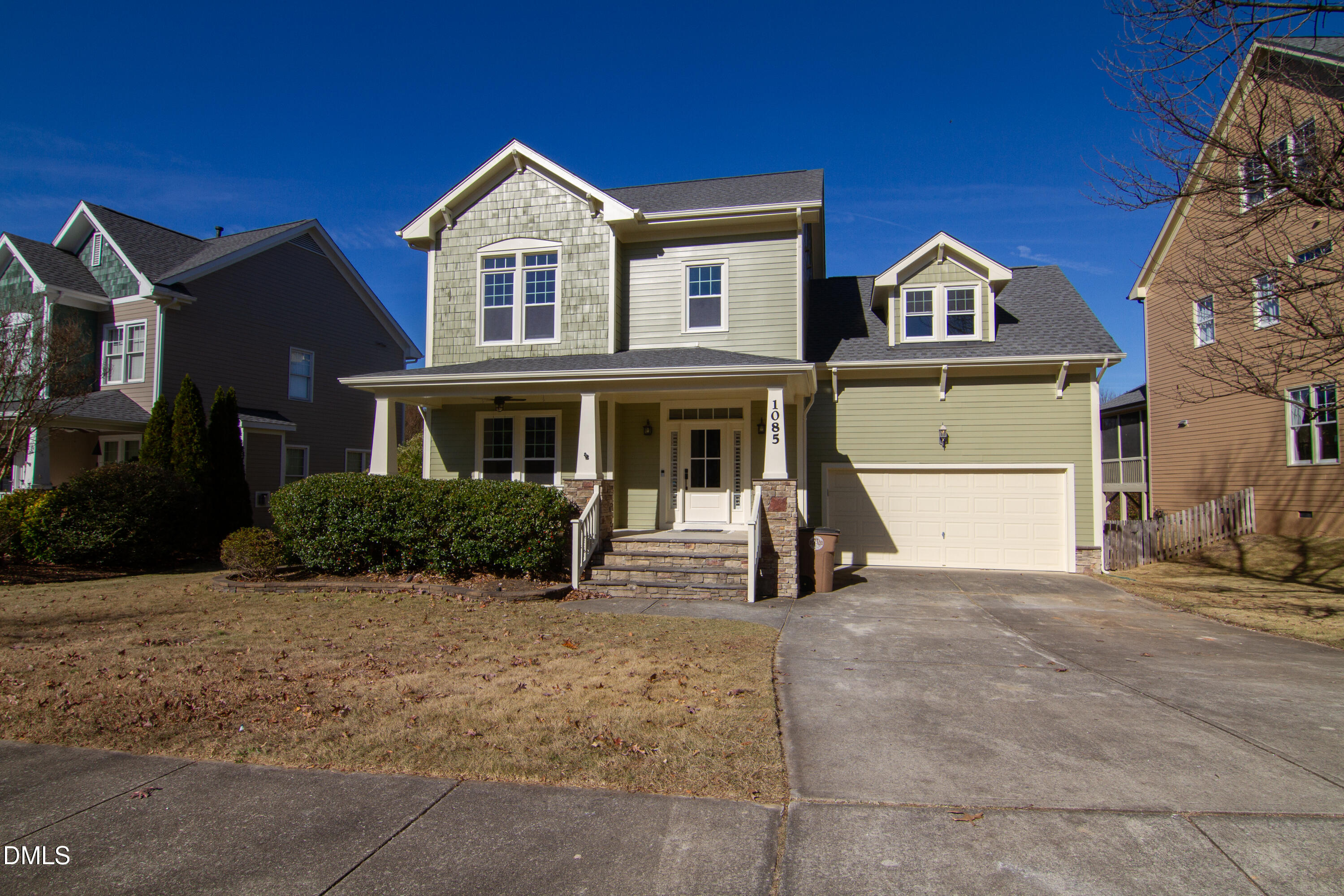 Two-story light-green house with a covered porch, stone supports, and an attached two-car garage.