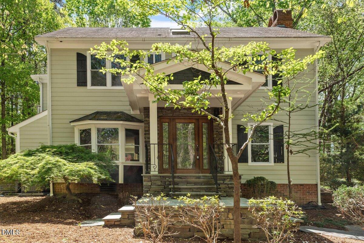 Light-sided home with a stone entry porch and bay window, framed by leafy trees.