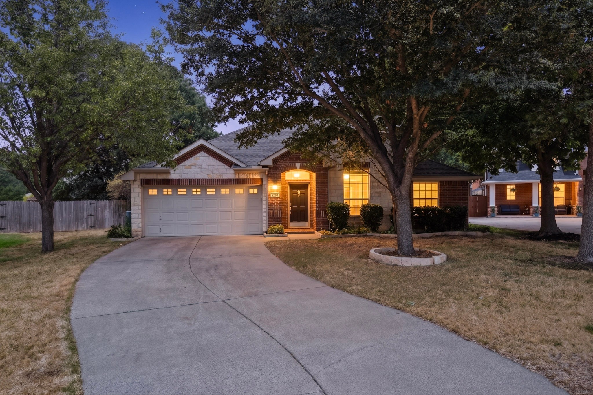 Single-story brick and stone house with a two-car garage and warmly lit windows beneath mature trees at dusk.