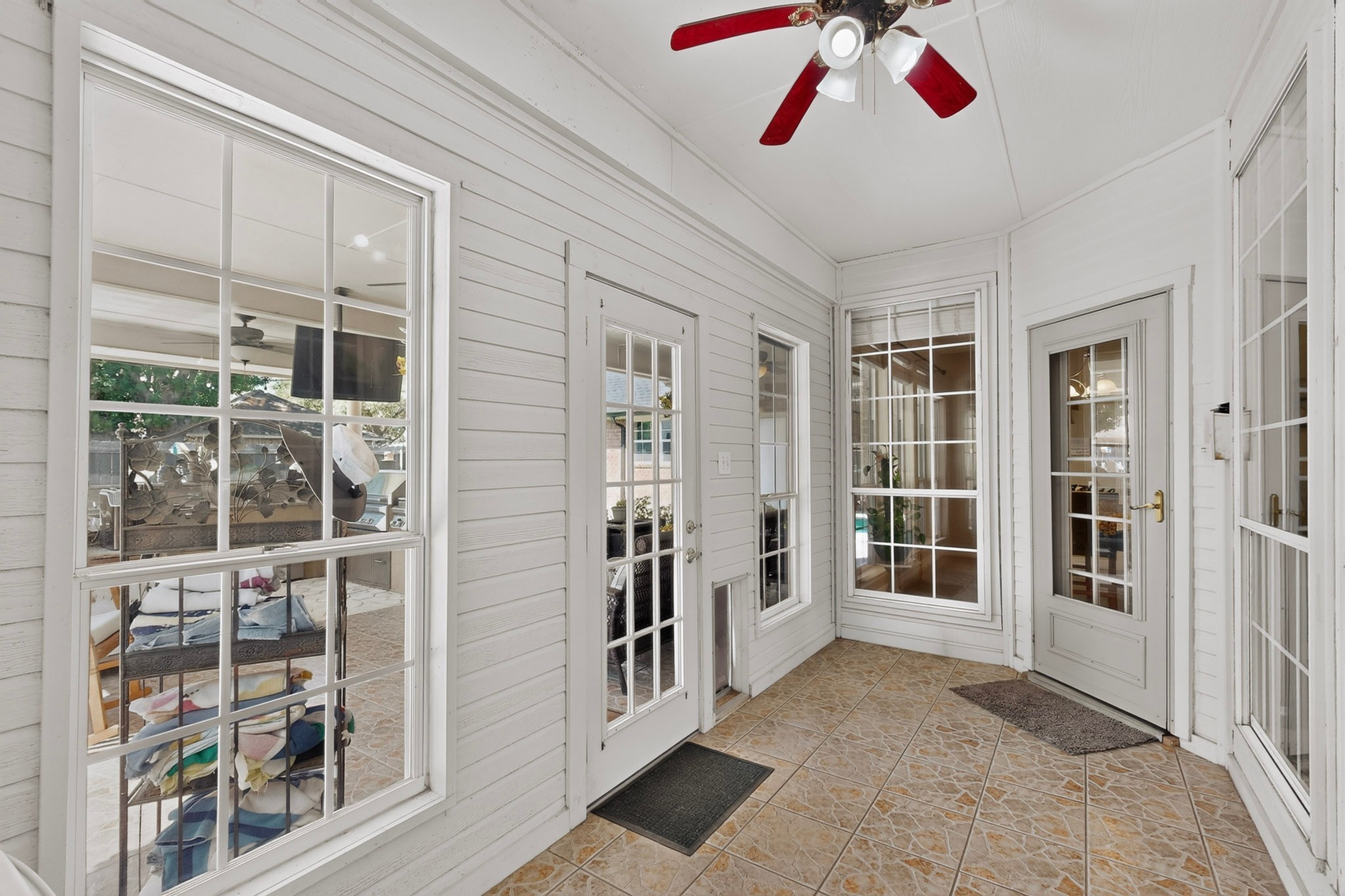 Enclosed sunroom with large windows, tan tile flooring, and a red-bladed ceiling fan.