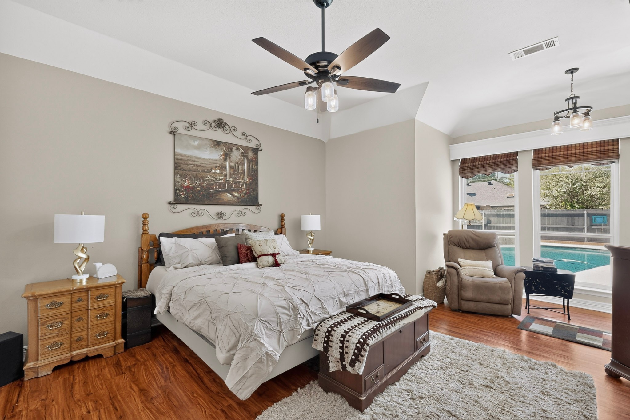 Furnished bedroom with vaulted ceiling and windows overlooking a backyard pool.