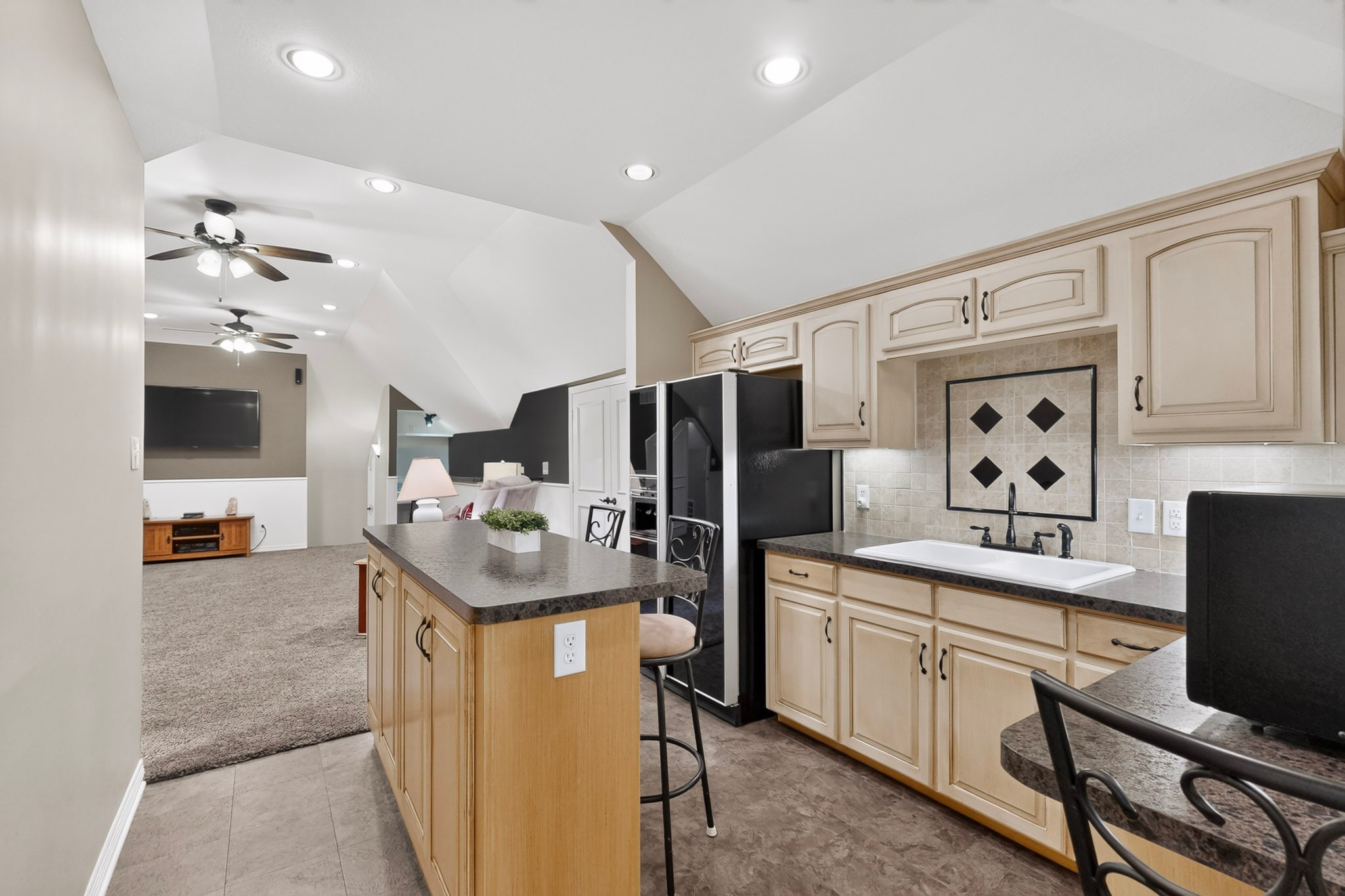 Kitchen with light wood cabinetry, dark counters, central island, and adjoining carpeted living area.