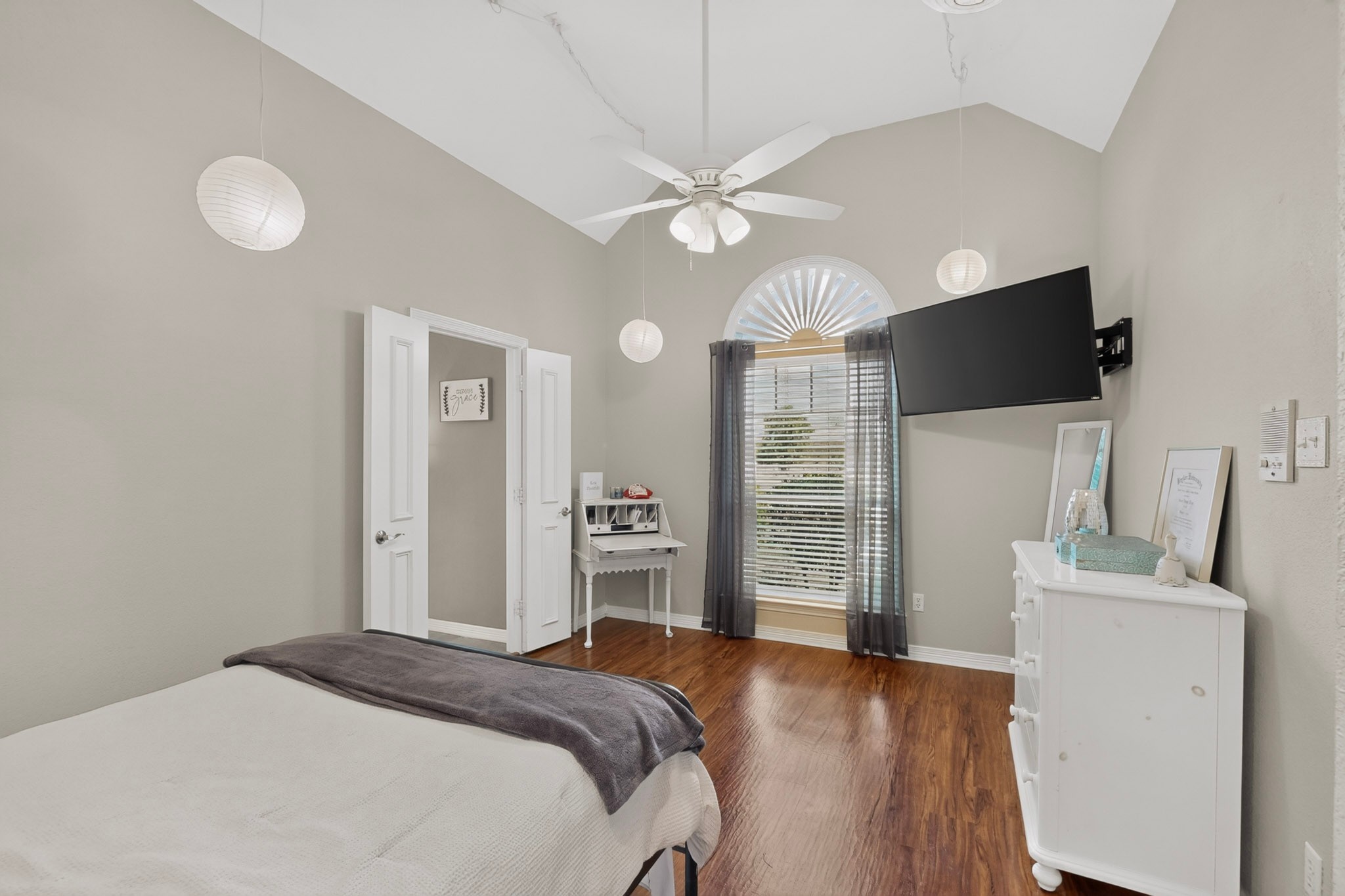 Bedroom with a vaulted ceiling, arched window, wall-mounted television, and polished hardwood floor.