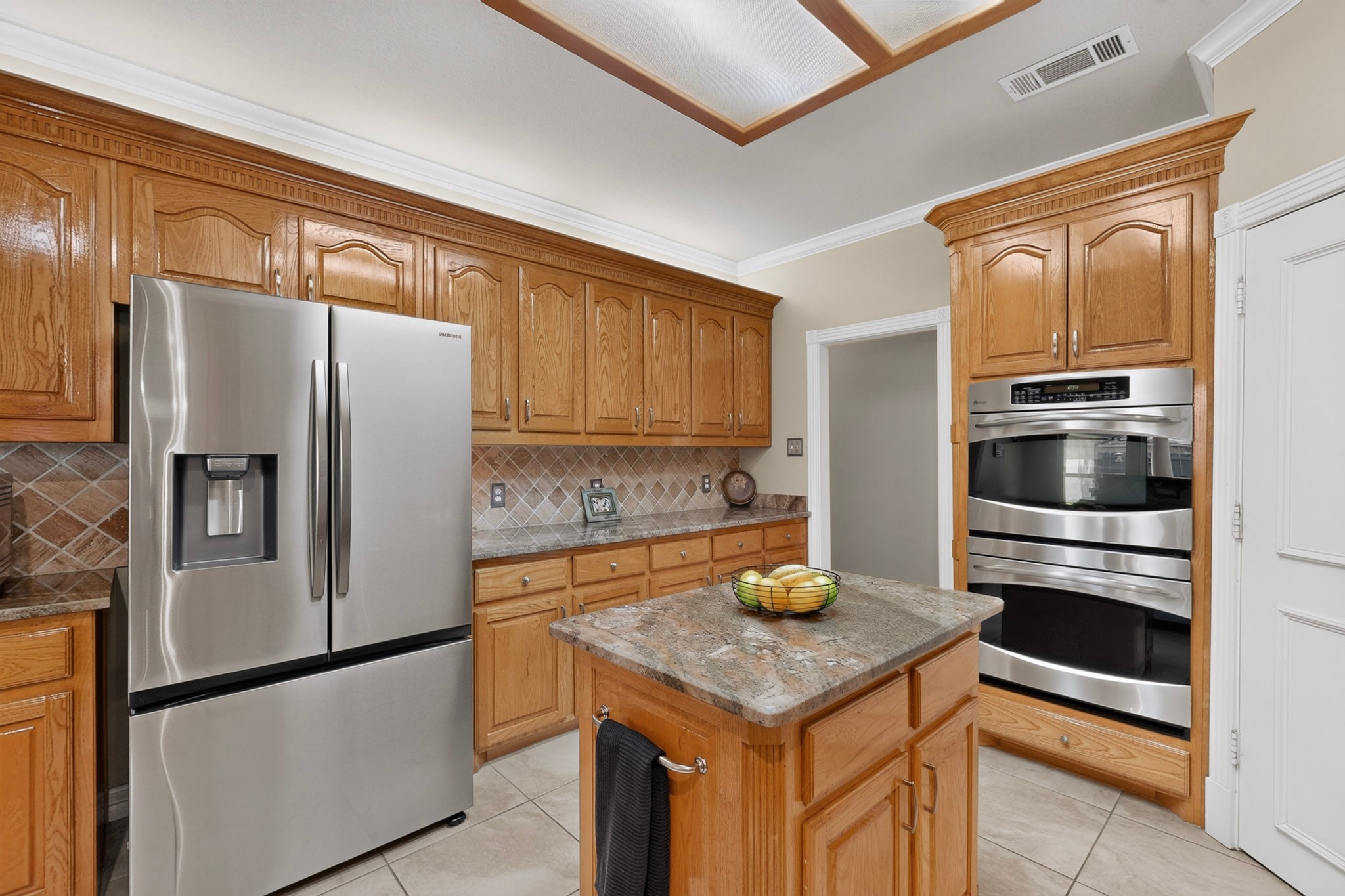 Kitchen with honey-colored wood cabinets, granite island, stainless refrigerator, and built-in double ovens.