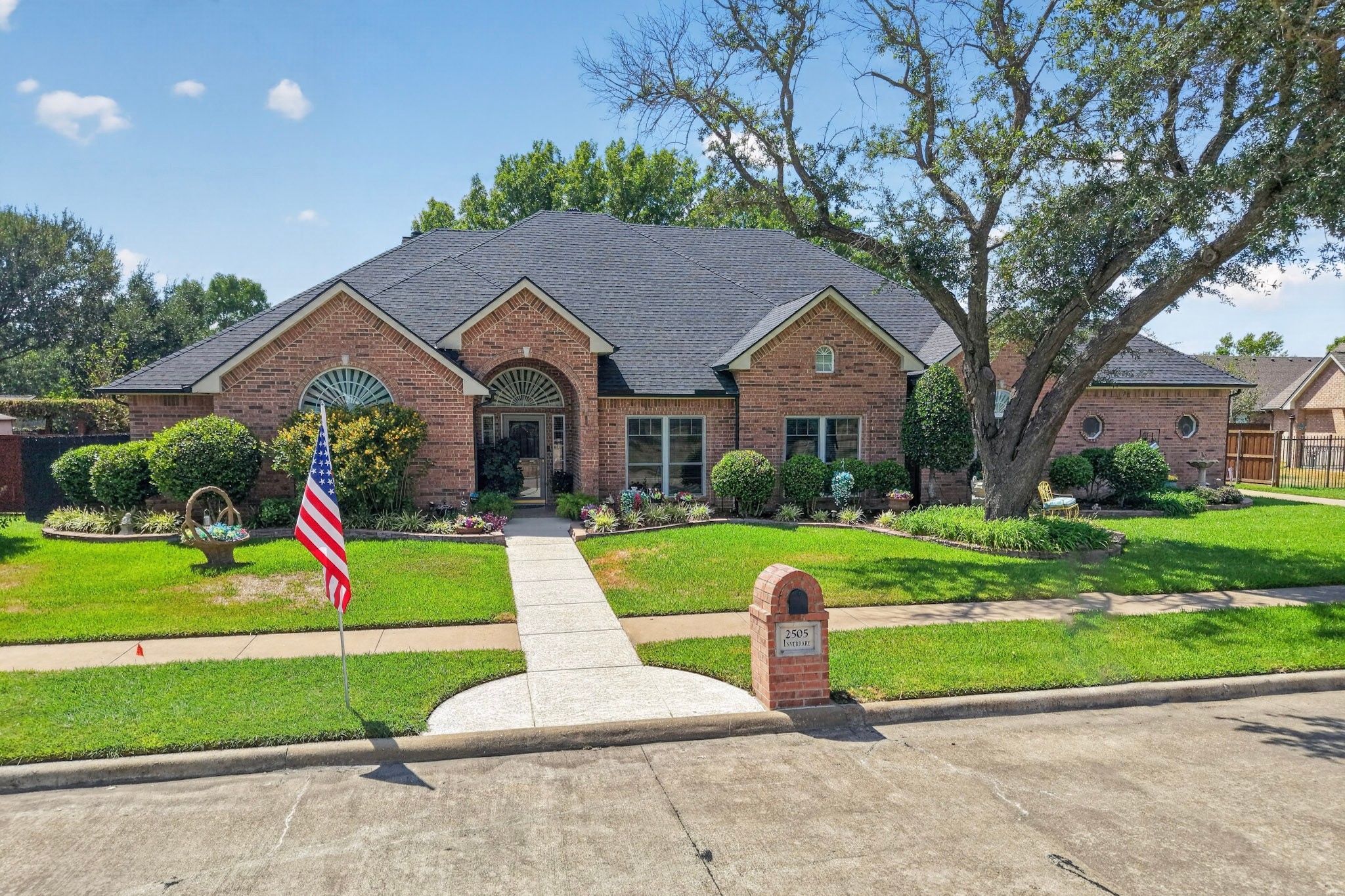 Red brick home with arched windows, landscaped lawn, large shade tree, and American flag.