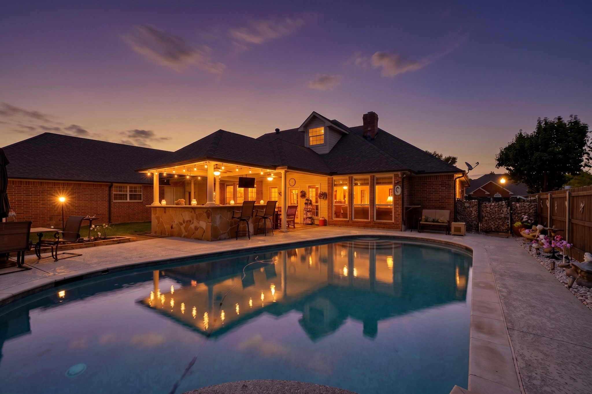 Backyard pool beside a brick home and illuminated covered patio at dusk.