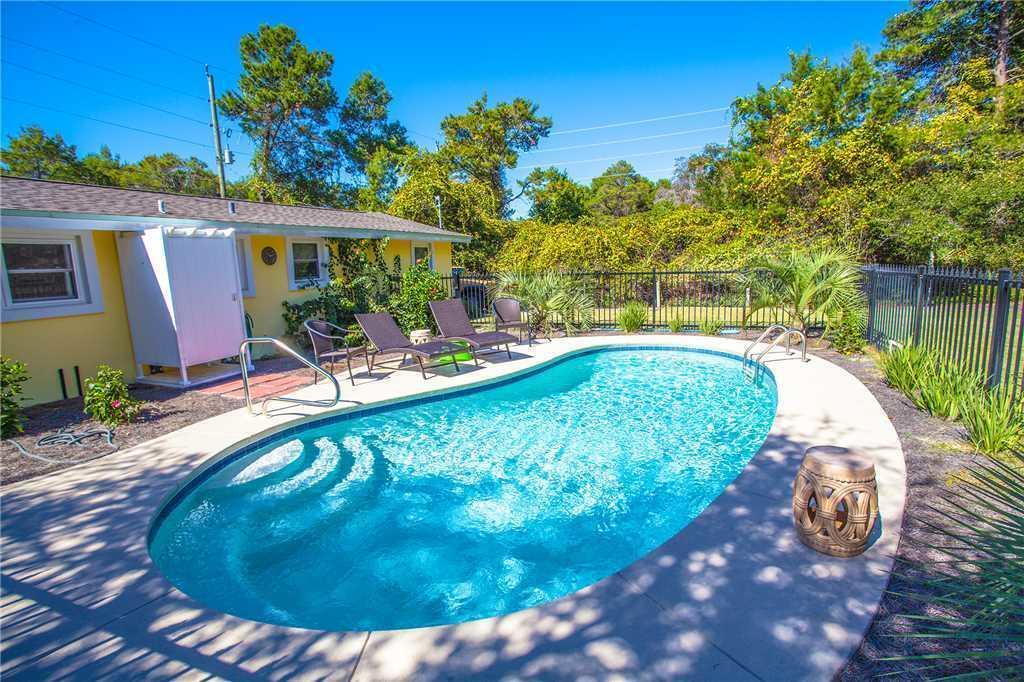 Curved backyard swimming pool beside a yellow house, with lounge chairs and a fenced, leafy yard.