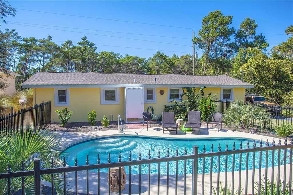 Yellow single-story house behind a fenced swimming pool, with patio chairs and tall trees.