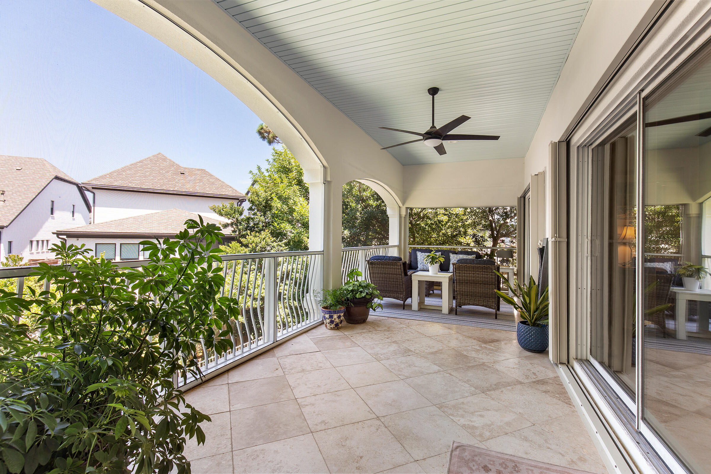 Covered tiled porch with ceiling fan, wicker seating, and open arches overlooking leafy surroundings.