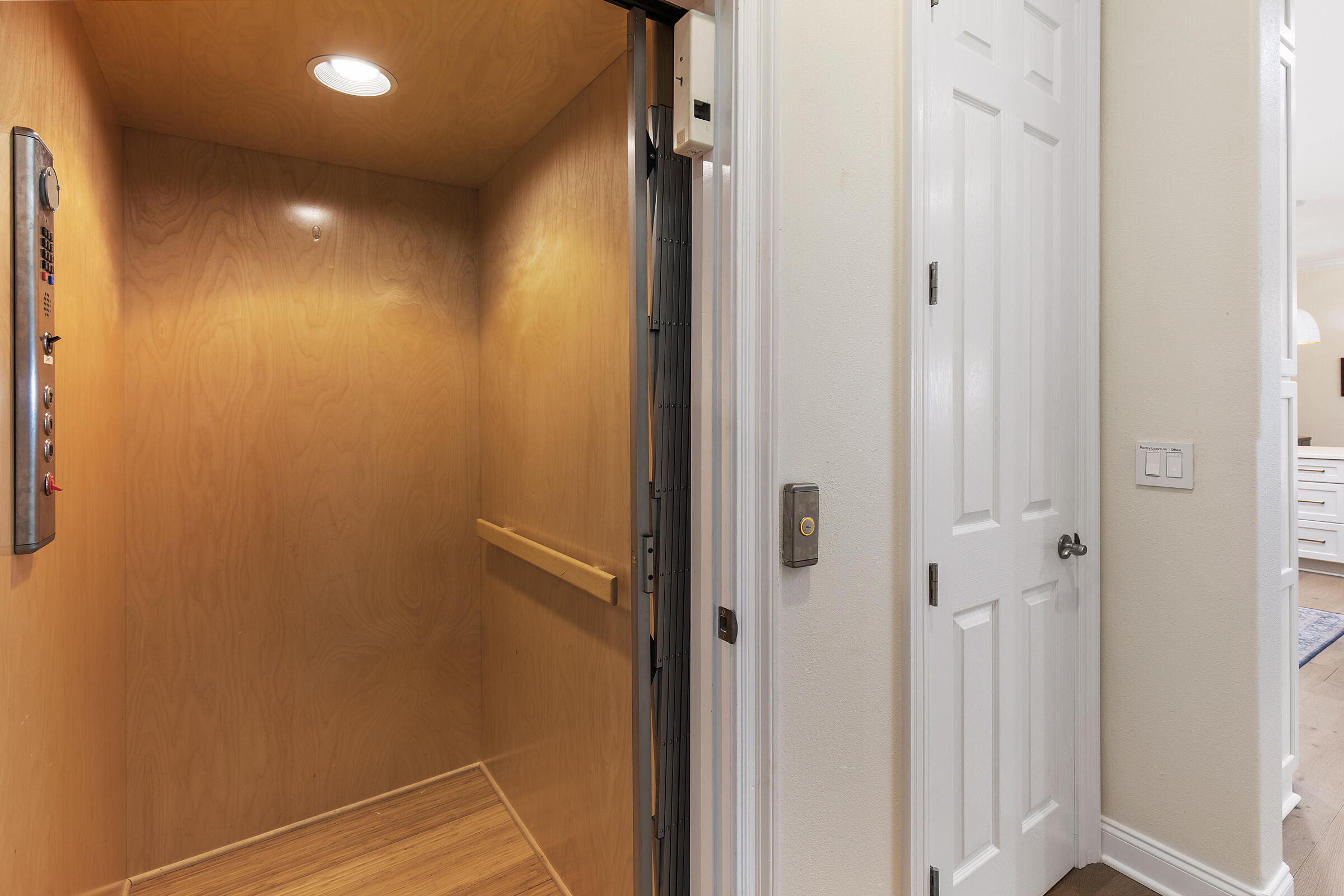 Wood-paneled residential elevator with a metal control panel beside a white hallway door.