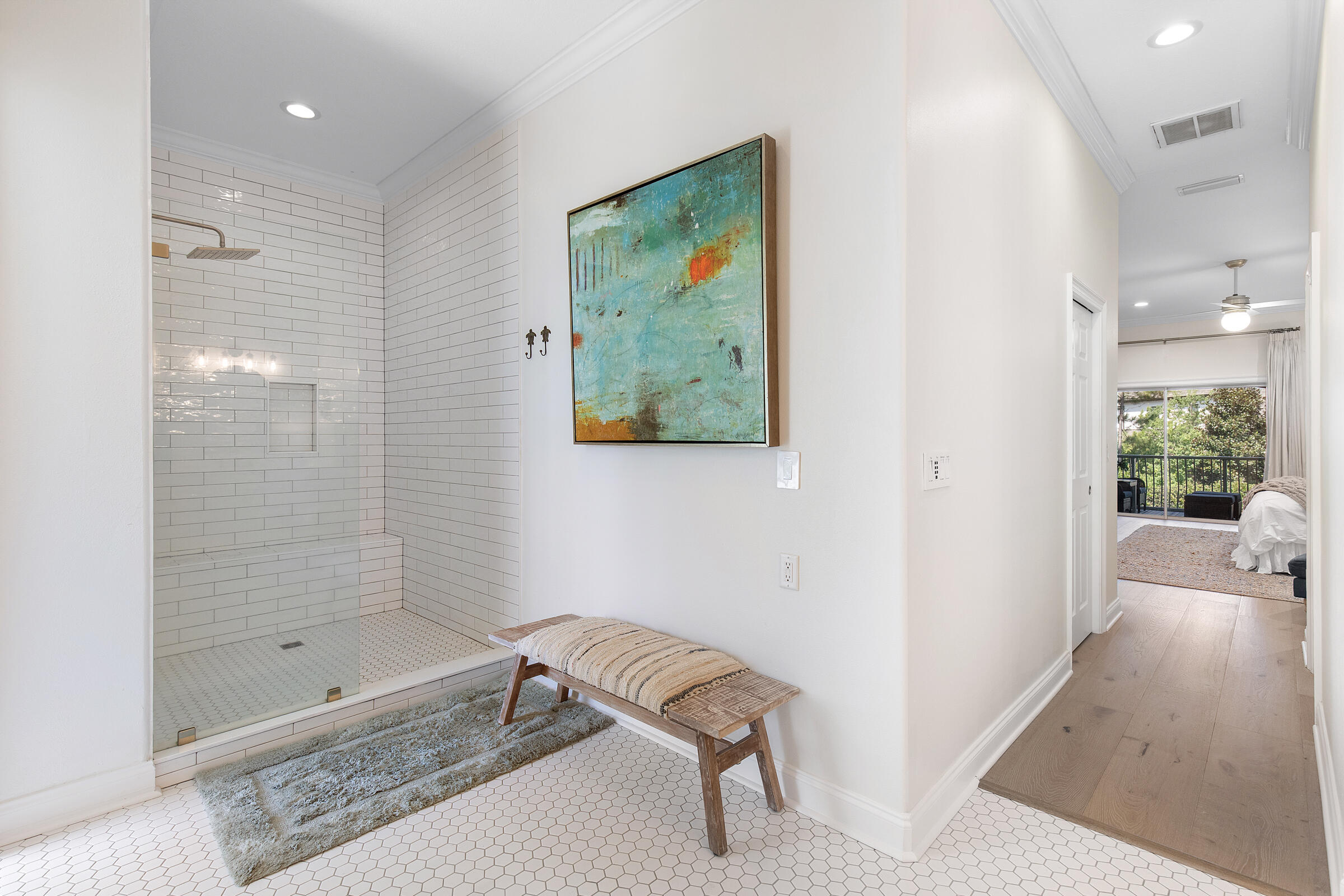 Bright bathroom with a tiled walk-in shower and woven bench, opening toward a bedroom and balcony.