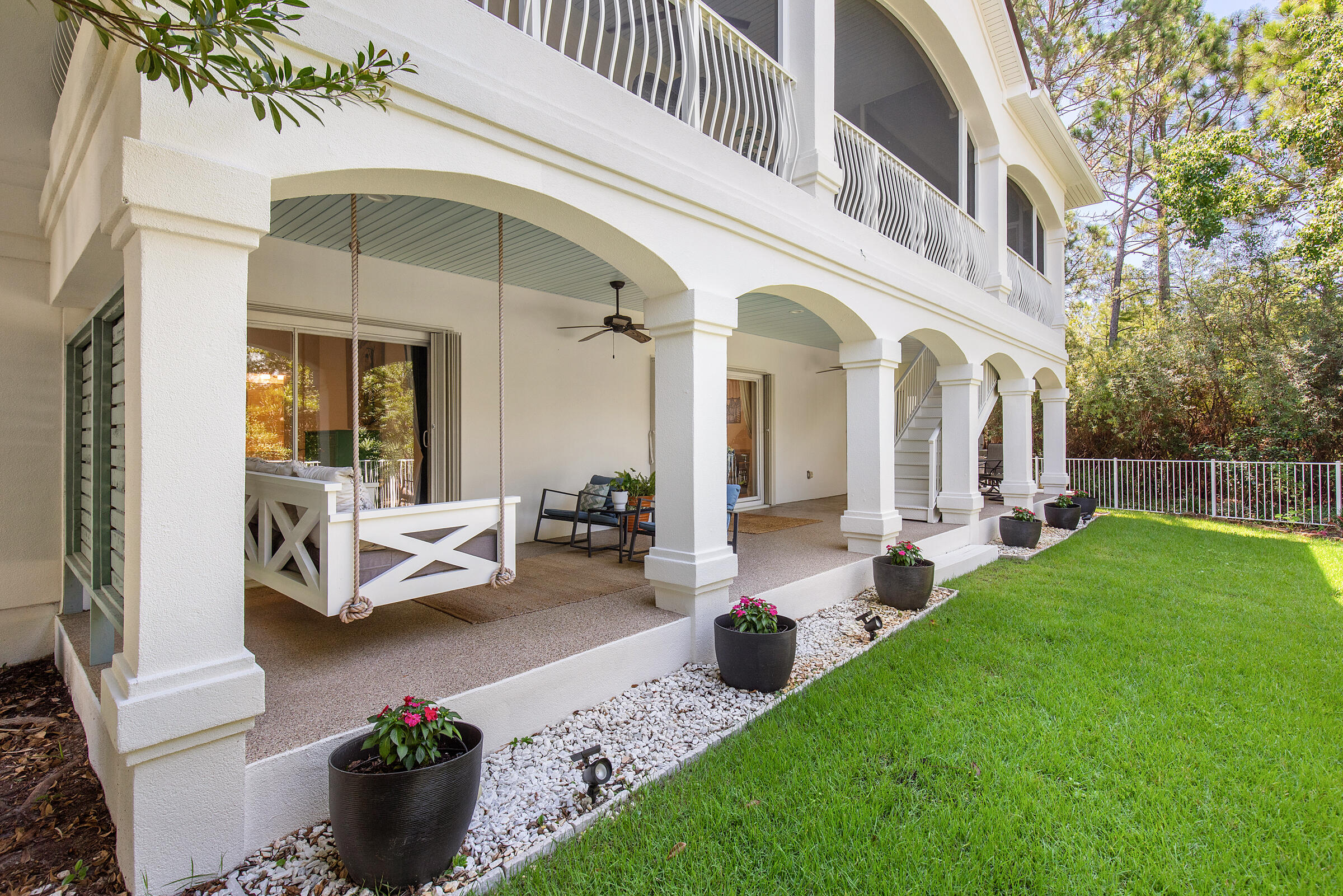White coastal home with an arched covered porch, hanging swing, upstairs balcony, and flower pots beside the lawn.