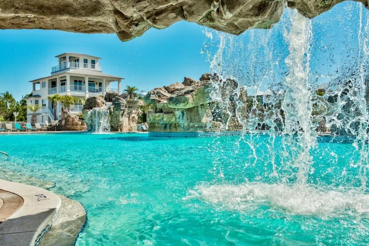 Turquoise pool with a rock waterfall and a white multi-story house in the background.